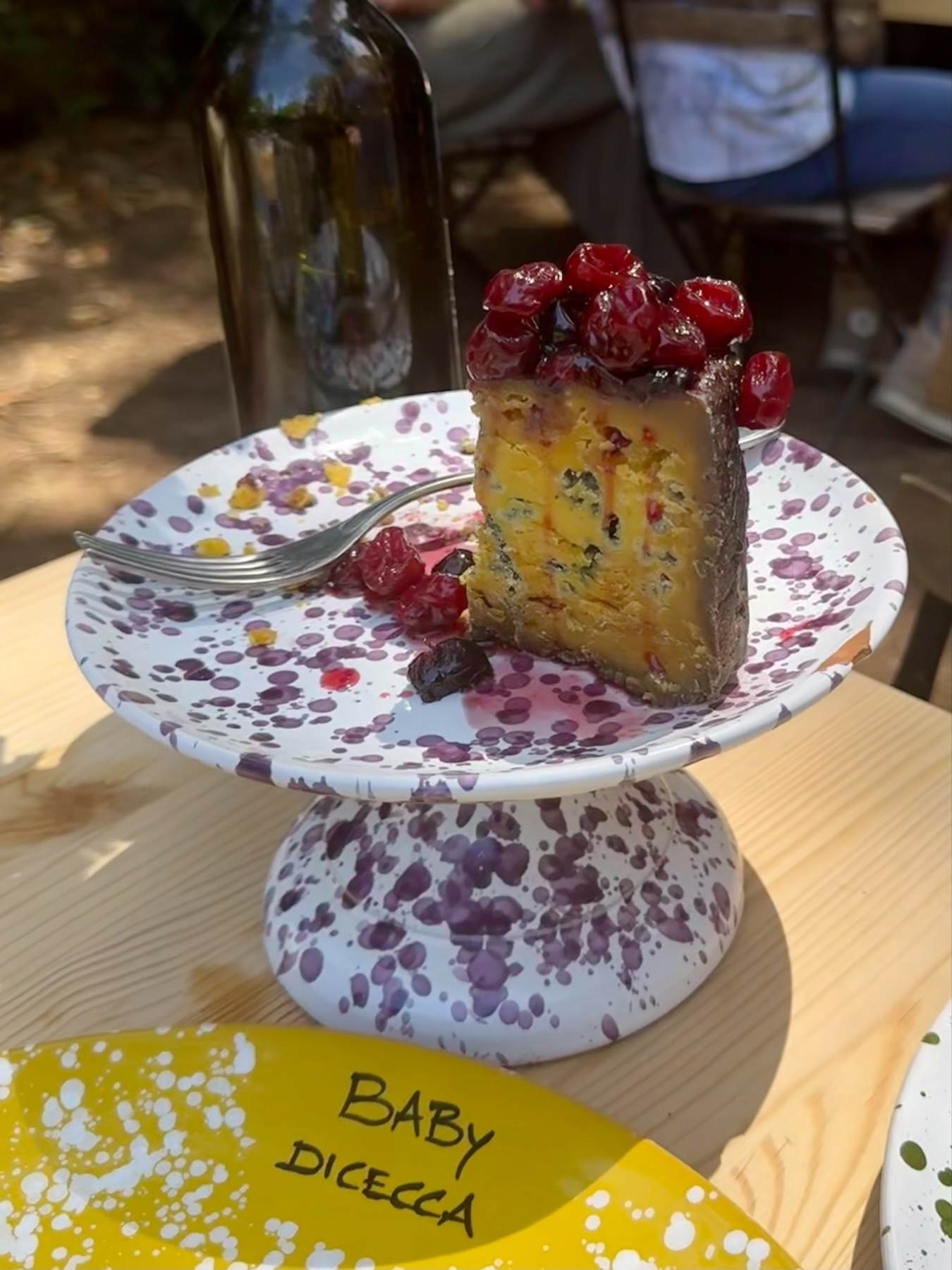 A slice of cake topped with berries is on a speckled cake stand, accompanied by a fork. There is a wine bottle in the background. In the foreground, a yellow plate labeled “BABY DICECCA” is visible. 