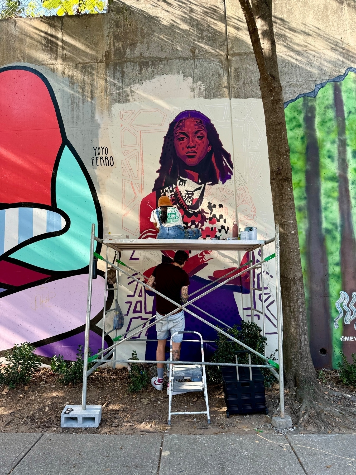 An artist, standing on a small step ladder and surrounded by scaffolding, is painting a colorful mural on a concrete wall. The mural features a detailed portrait of a person with long hair and distinctive facial markings.