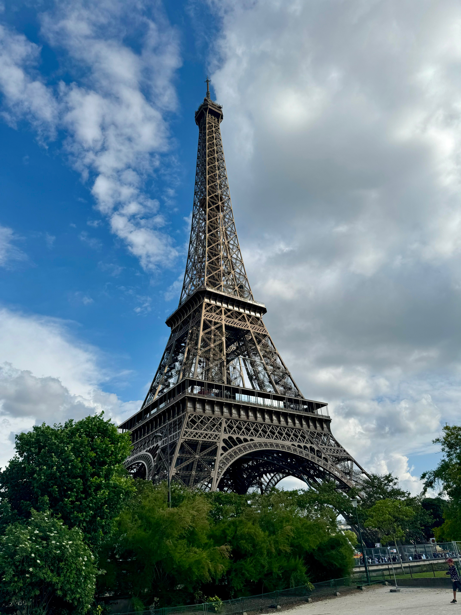 The Eiffel Tower stands tall against a blue sky with clouds, surrounded by green foliage. A person is visible at the bottom right corner, appearing small in comparison to the towering structure.