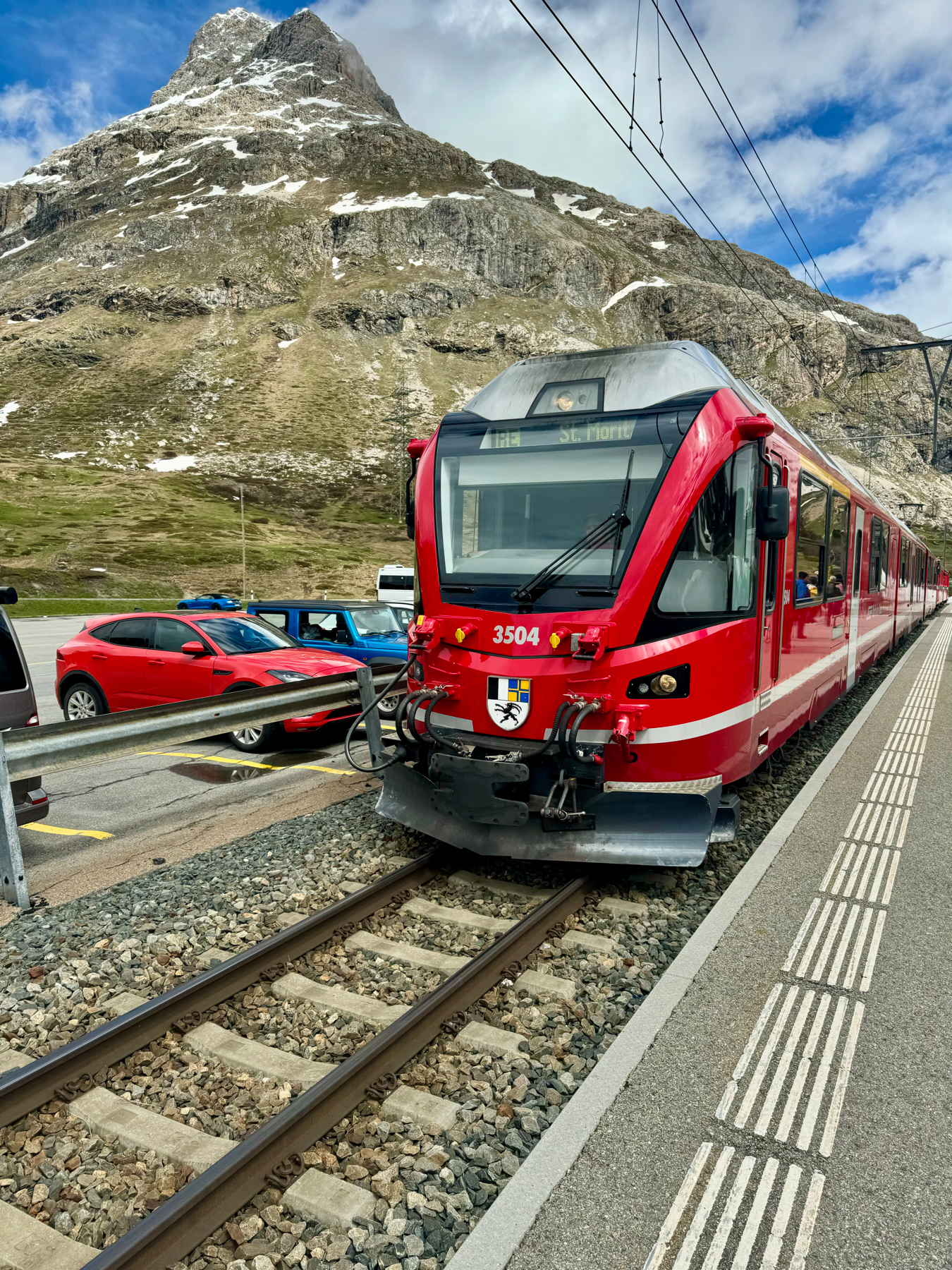 A red train with the destination sign “St. Moritz” on the front, traveling on tracks surrounded by cars with a rocky, snow-patched mountain in the background.