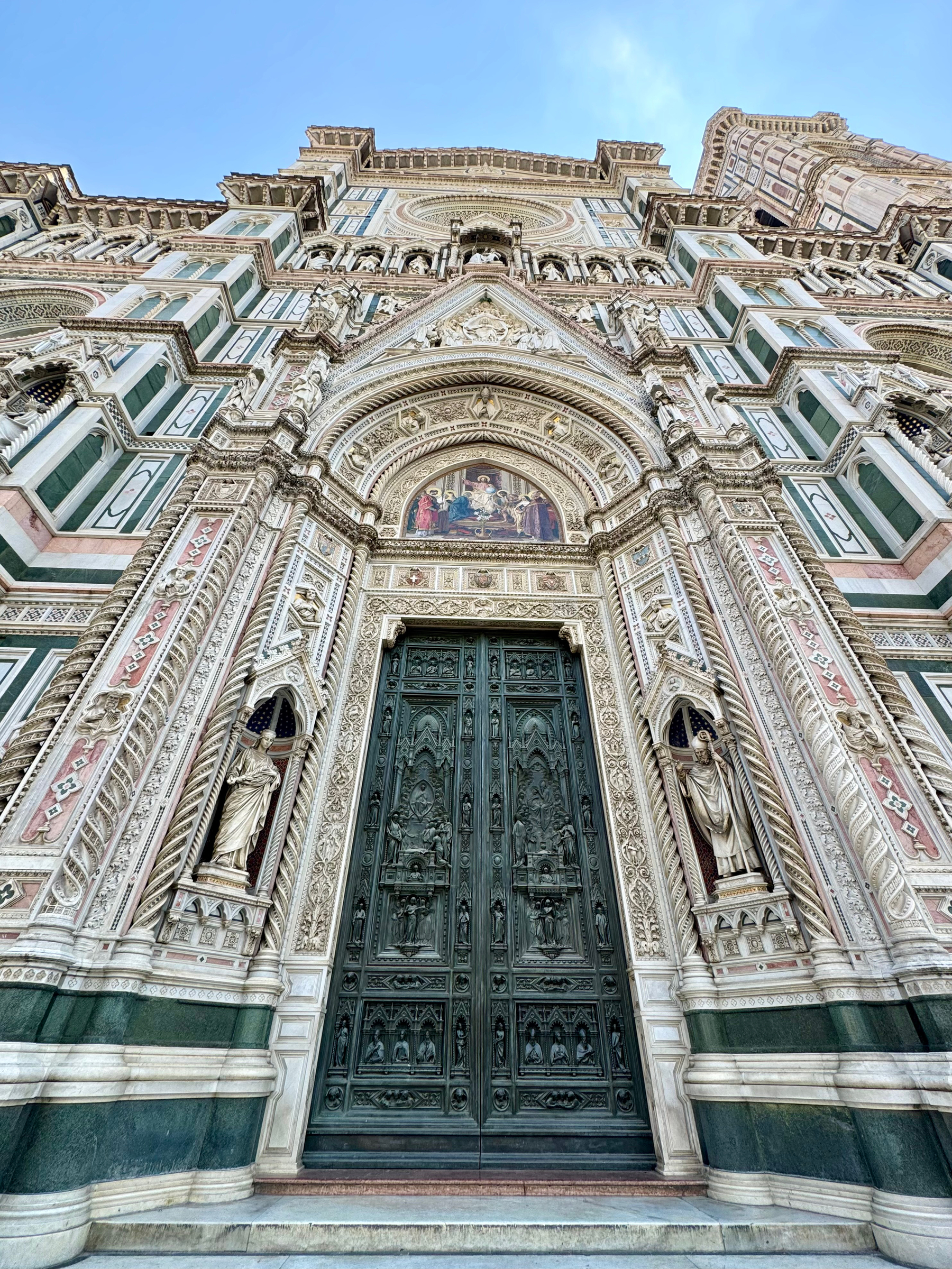 This image shows the intricate and ornate facade of Florence Cathedral, also known as the Cathedral of Santa Maria del Fiore, in Florence, Italy. The focus is on the central main doors, which are richly decorated with religious carvings and sculptures. 