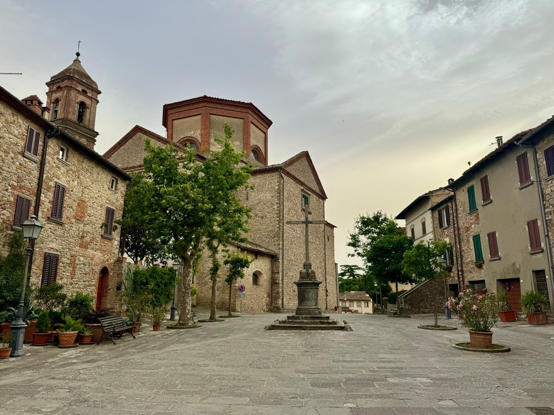 A quaint, empty village square with stone buildings, a church with a bell tower, and a large cross monument in the center. The scene features cobblestone pavement, potted plants, trees, and benches, all under a cloudy sky.
