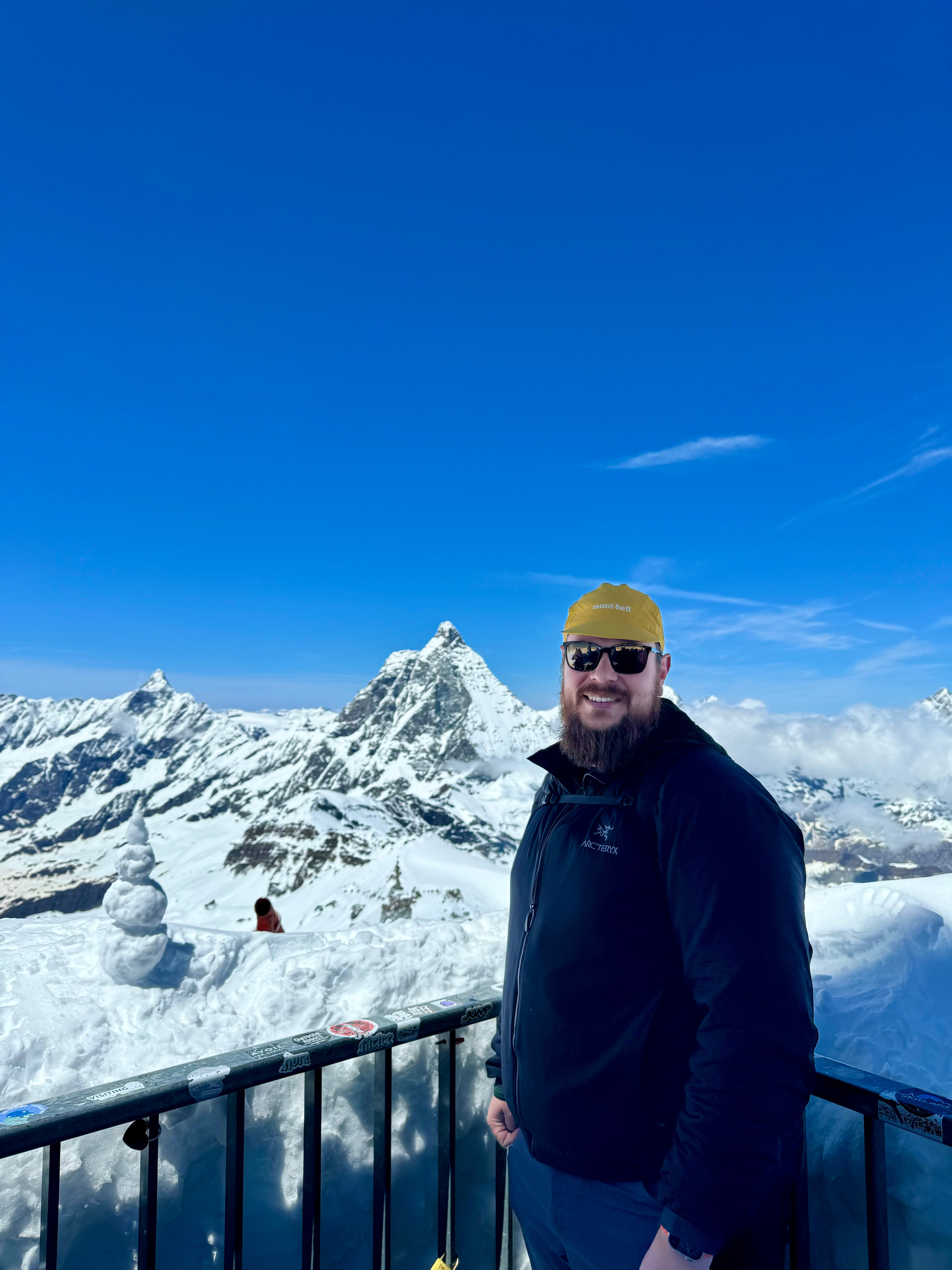 A man with a beard wearing a black jacket and a yellow hat stands smiling in front of a snow-covered mountain range under a clear blue sky.