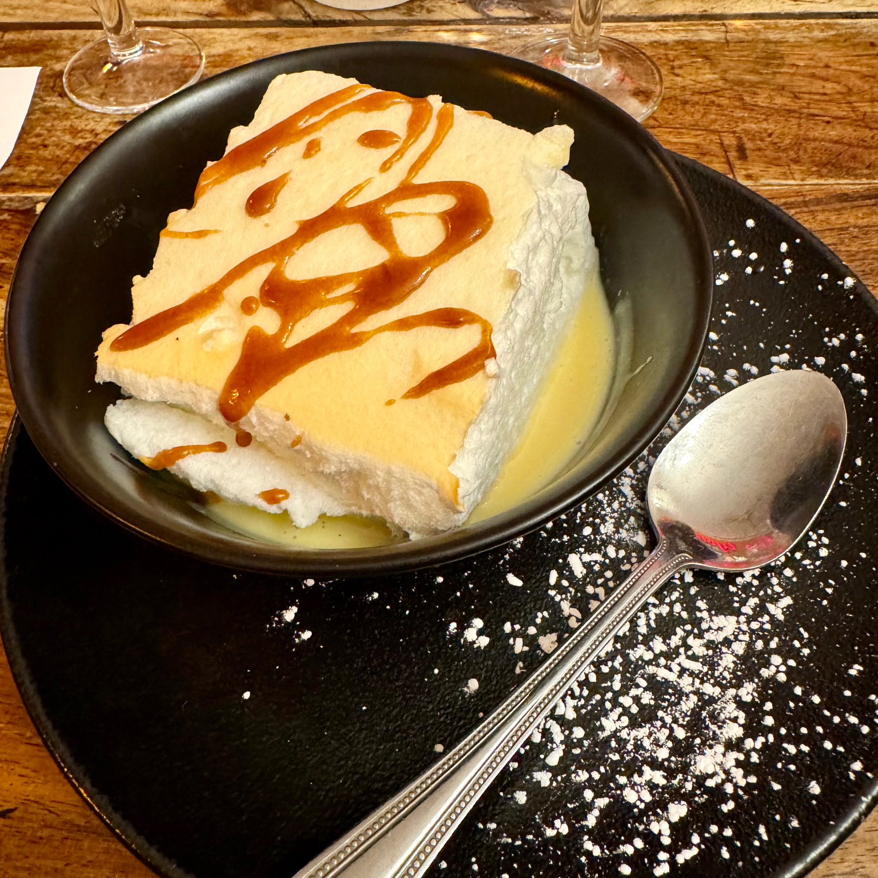 A slice of meringue in a pool of custard with caramel sauce and powdered sugar on a black plate with a spoon, on a wooden table.