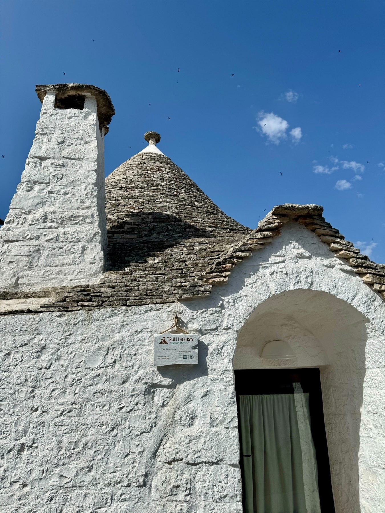 A close-up view of a traditional trullo house with whitewashed stone walls and a conical roof made of stone slabs. The house features a chimney and an arched entrance covered with a curtain. 