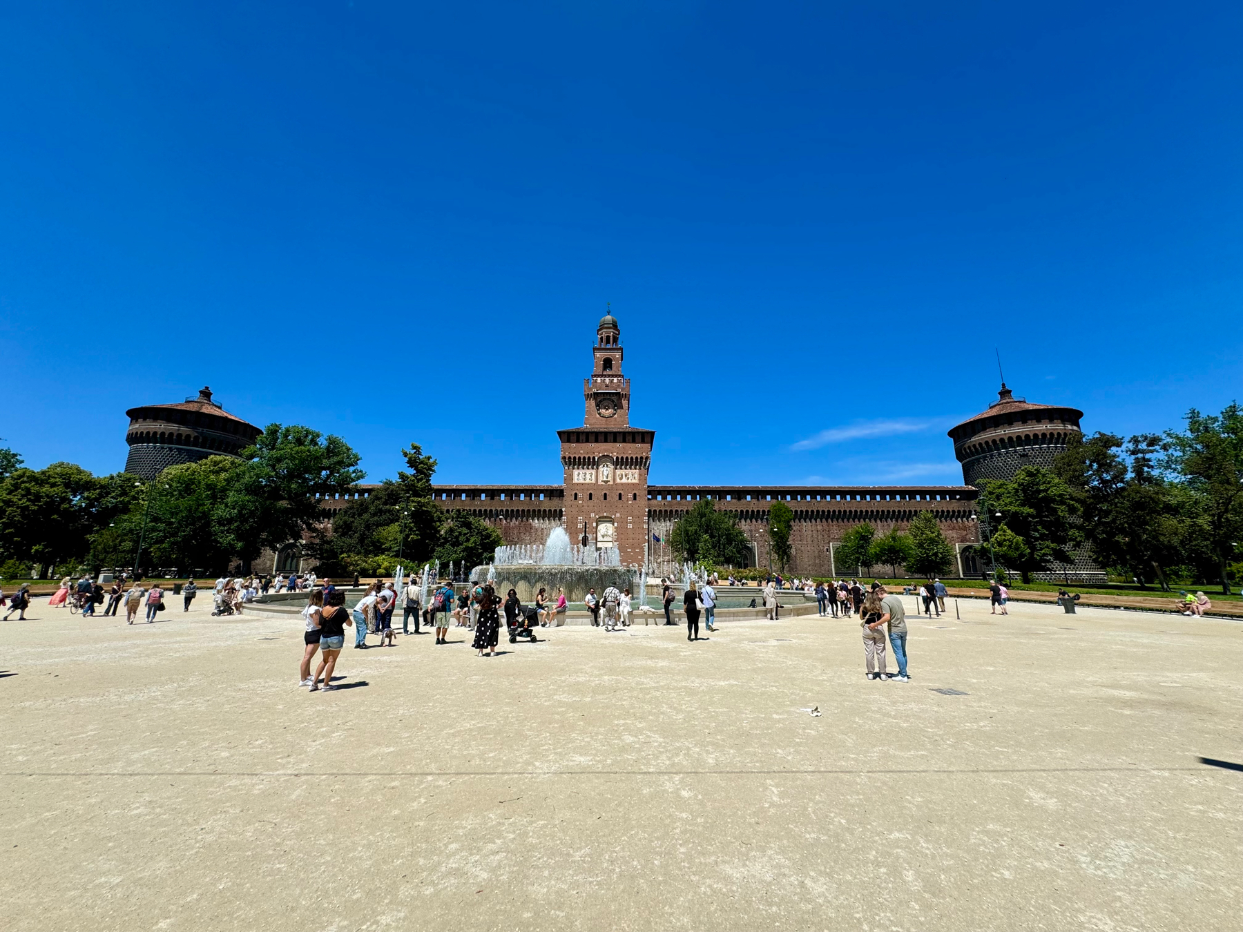 Image of Sforza Castle (Castello Sforzesco) in Milan, Italy, on a clear day. The castle’s central tower and side towers are prominent. There are people walking and gathering near the fountain in the foreground.