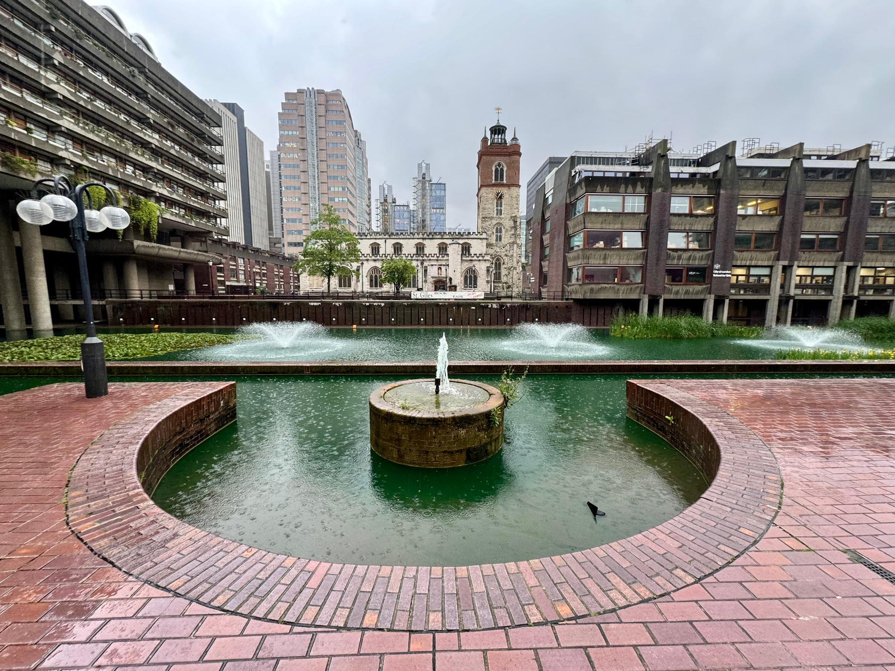 A fountain in a pond, surrounded by brick pavement with brutalist architecture and modern buildings in the background, under a rainy sky.