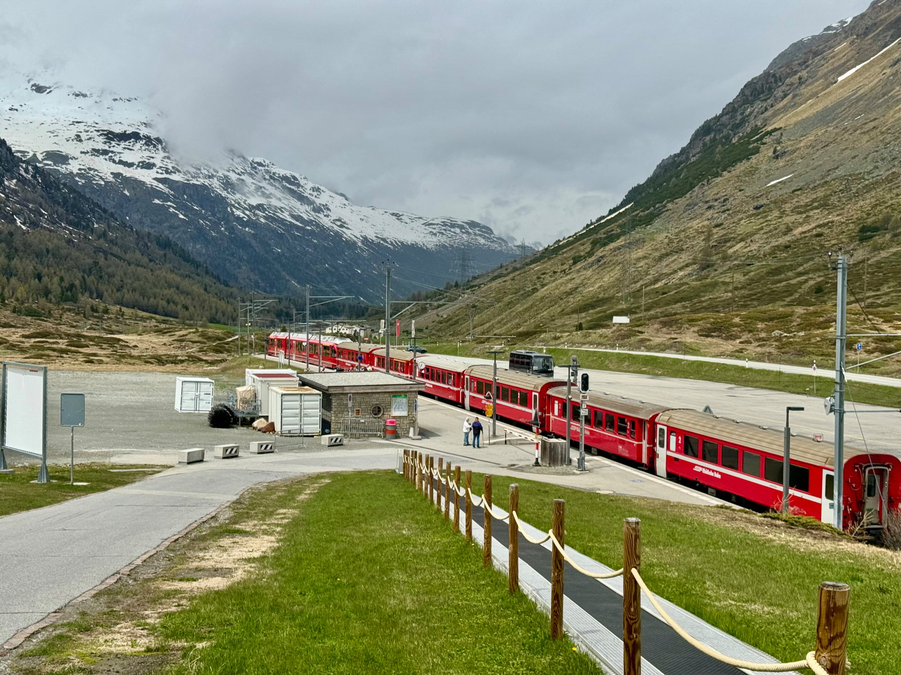 A red passenger train stationed at a platform amidst a mountainous landscape with snow-capped peaks and a grassy area in the foreground.