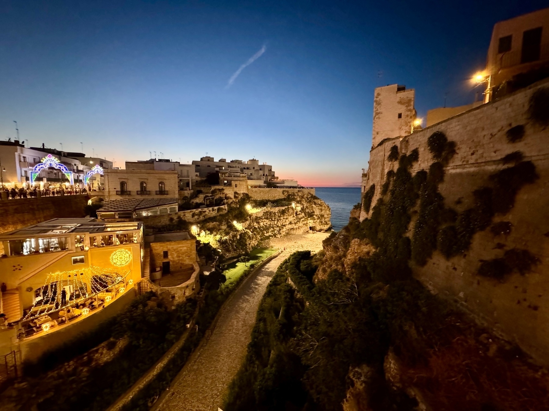 Dusk view of a coastal town featuring illuminated buildings, a lit pathway, and a rocky cove leading to the sea. The sky is a gradient from dark blue to a faint orange horizon, and the architecture showcases a blend of modern and old structures. 