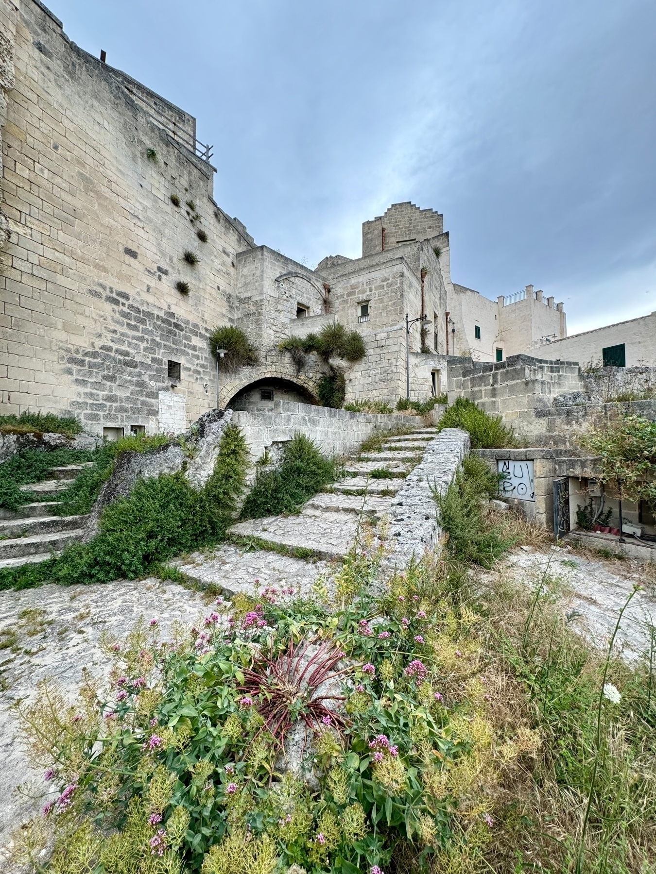 A historic stone structure with a weathered, rustic appearance. The image shows a series of stone steps, lush greenery, and flowering plants in the foreground. Buildings with arched doorways and small windows are present, and the stone walls have plants growing. 