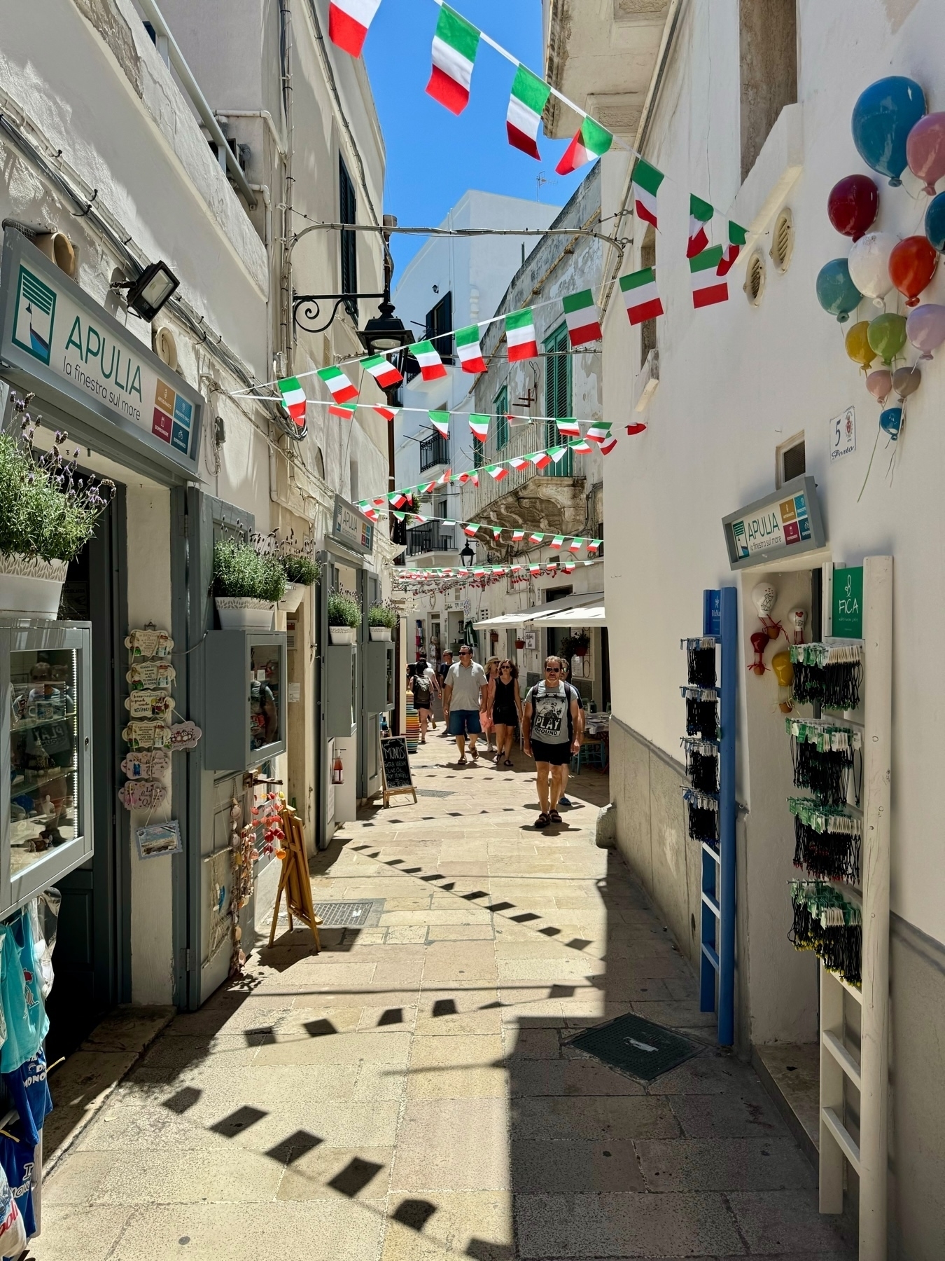 Narrow pedestrian street adorned with Italian flag bunting. Shops on both sides display various items, including signs for “APULIA.” People stroll along the cobblestone path under a clear blue sky.