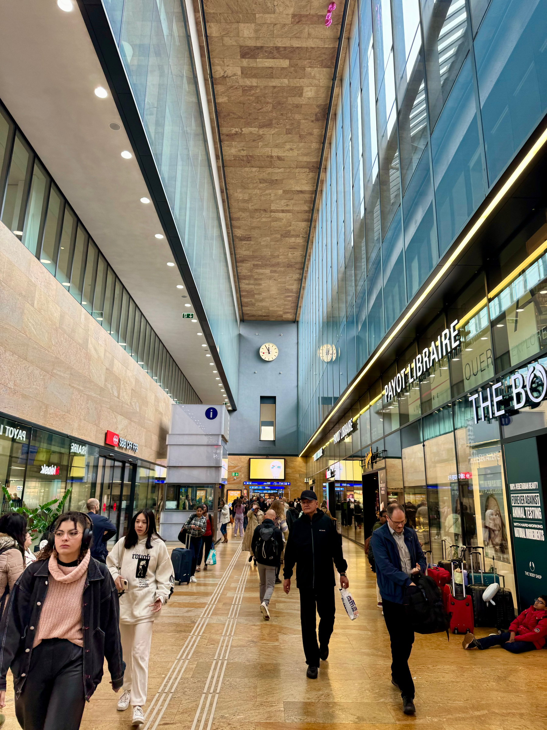 Indoor corridor in a modern building with people walking, shops on the right, decorative wood paneling on the ceiling, glass facade on the left, and hanging clocks.