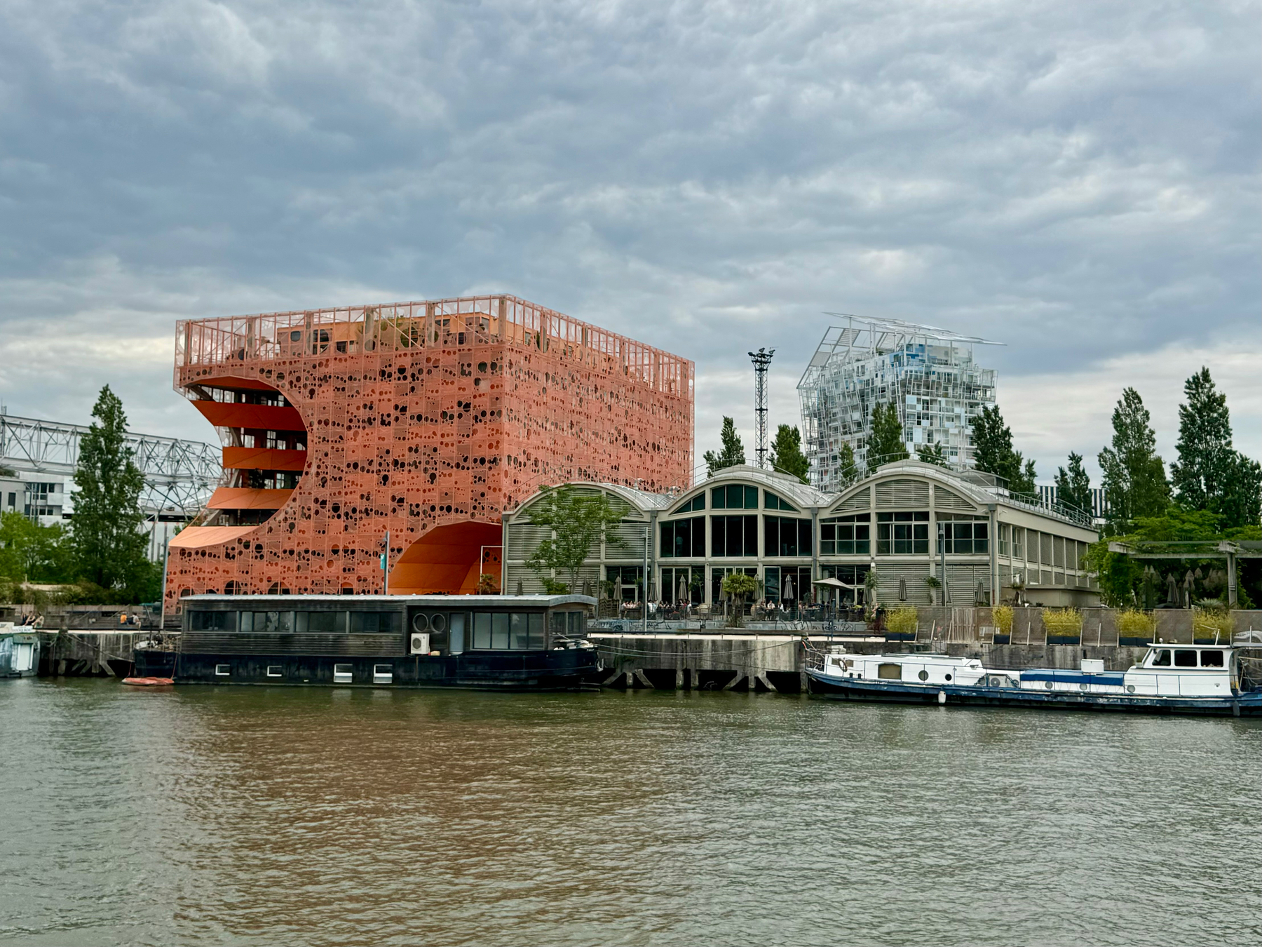 The image shows a riverside view with modern buildings, including a distinctive orange building with a unique perforated facade and a spiral staircase on its exterior. Behind it is a building with an intricate geometric glass structure on its top. There are boats moored. 