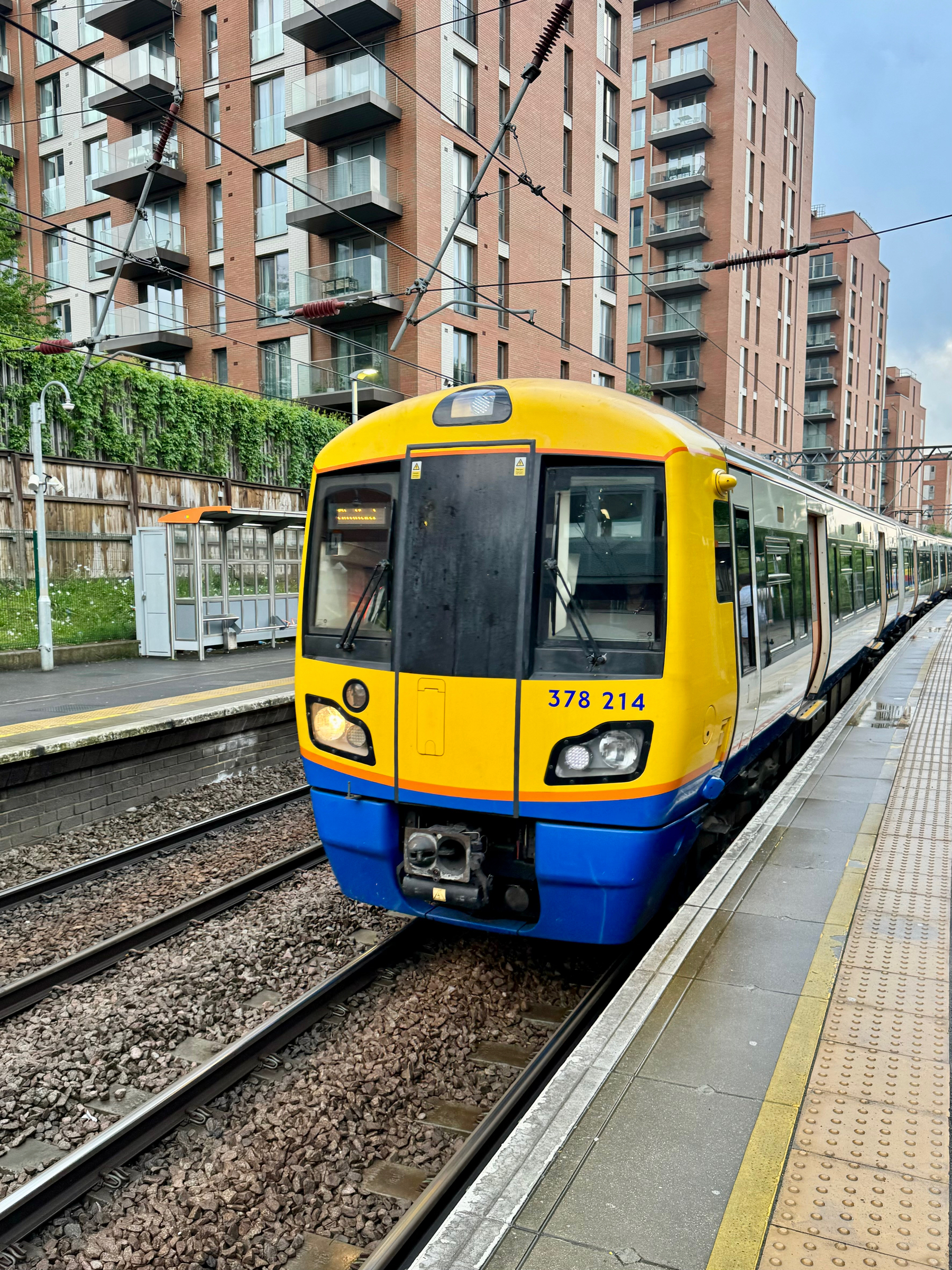 A yellow and blue Overground train, numbered 378 214, at a platform with apartment buildings in the background.