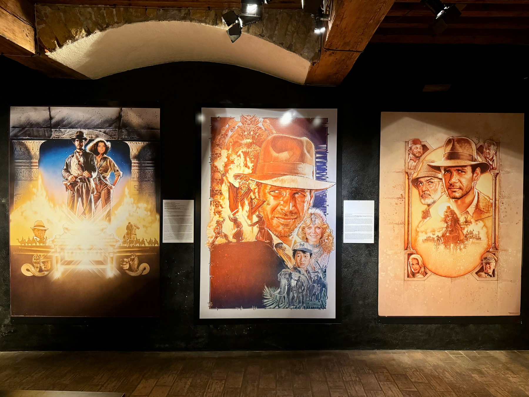 Three framed movie posters depicting the original Indiana Jones films, displayed in an exhibition with spot lighting and wooden beams in the background.