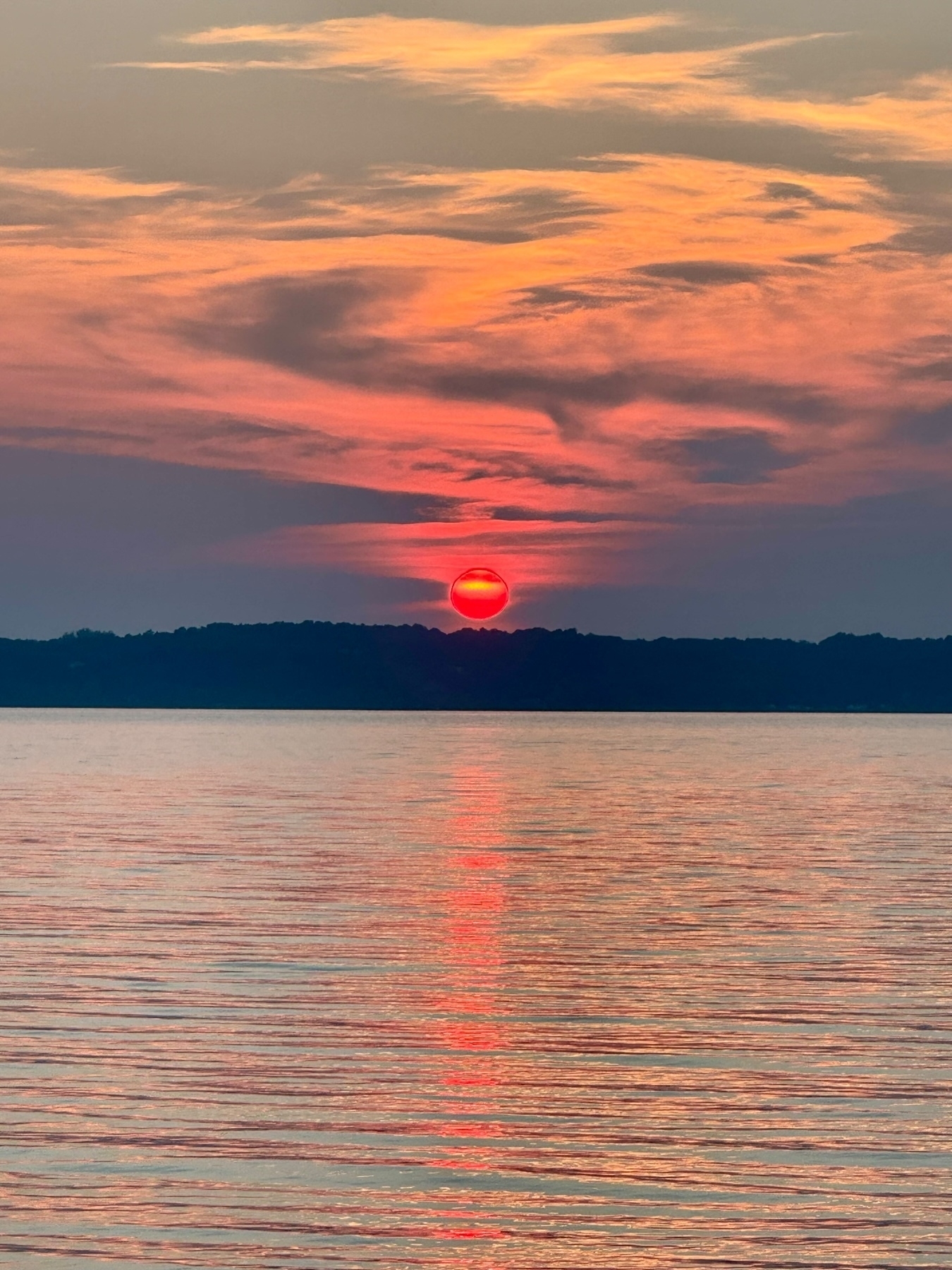 A vivid sunset with a bright red sun setting over a distant dark shoreline. Above, the sky is painted with shades of orange, pink, and purple clouds. The reflection of the sun creates a vertical red streak on the calm water below.