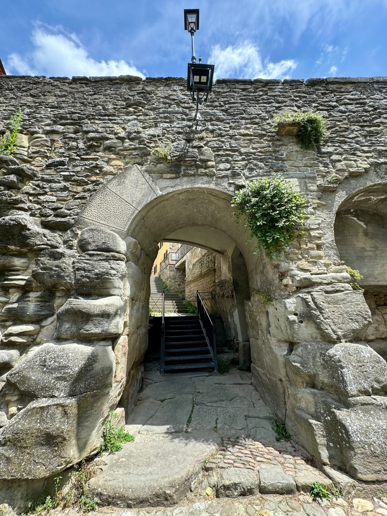 A historic stone archway with a lamp post on top. The arch leads to a staircase with metal railings, ascending towards buildings in the background. Vegetation grows through the stonework at various points, and cobblestones line the pathway under. 