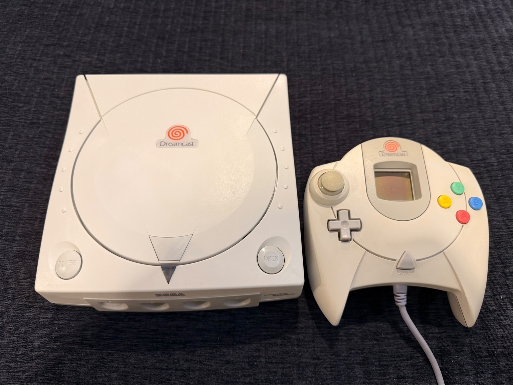 A Sega Dreamcast console and its controller on a dark surface. The console is white with the Dreamcast logo, and the controller features a joystick, a D-pad, and four colored buttons.