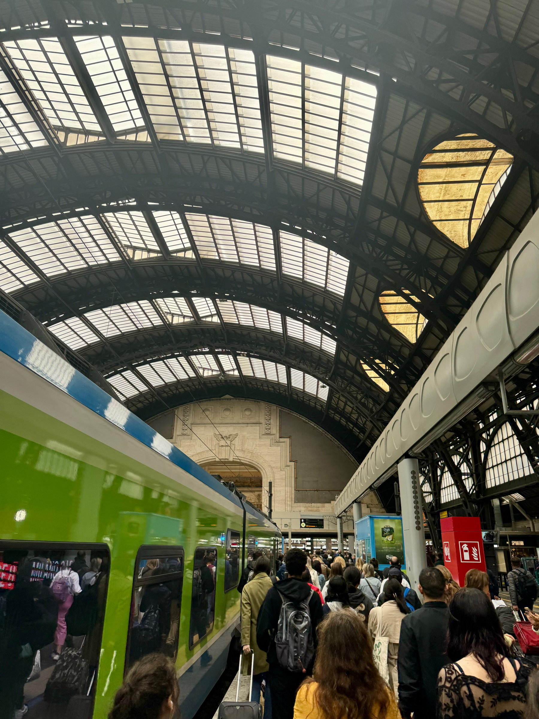 Crowded train station with passengers waiting on the platform. An electric train with green and blue coloring is parked. The station features a high, arched glass roof and intricate architectural details on the wall. 