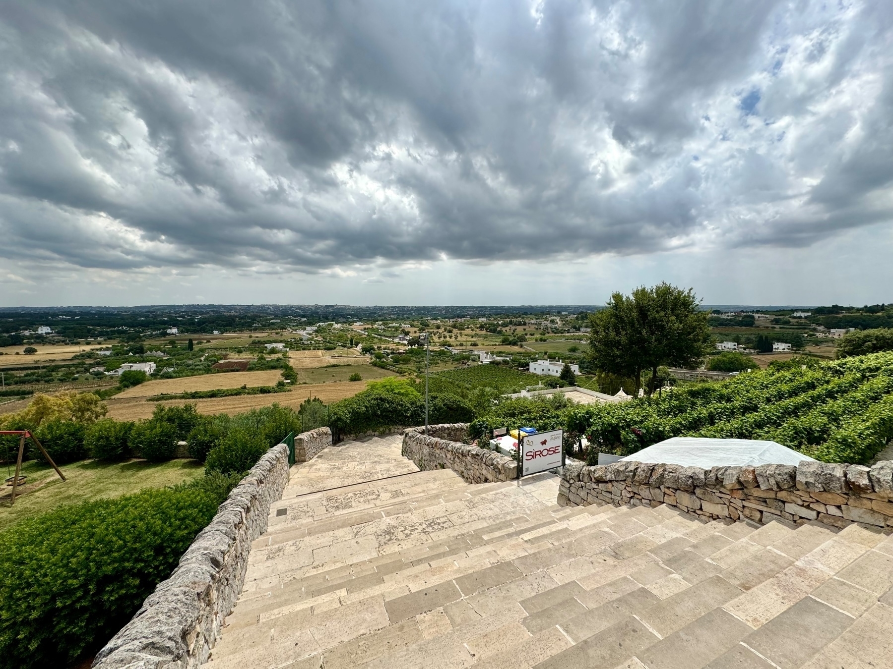 A panoramic view of a countryside landscape taken from a stone staircase. The image features cloudy skies, green vineyards, farmland, and scattered buildings surrounded by vegetation. A sign reading “SIROSE” is visible near the bottom of the stairs.