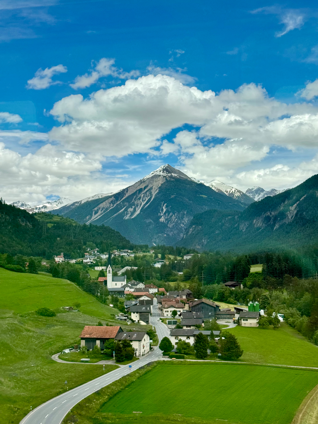 A picturesque village nestled at the base of a mountain range with a prominent peak against a blue sky with fluffy clouds. A road winds through vibrant green fields leading towards the village center, characterized by traditional buildings and a church with a tall steeple.