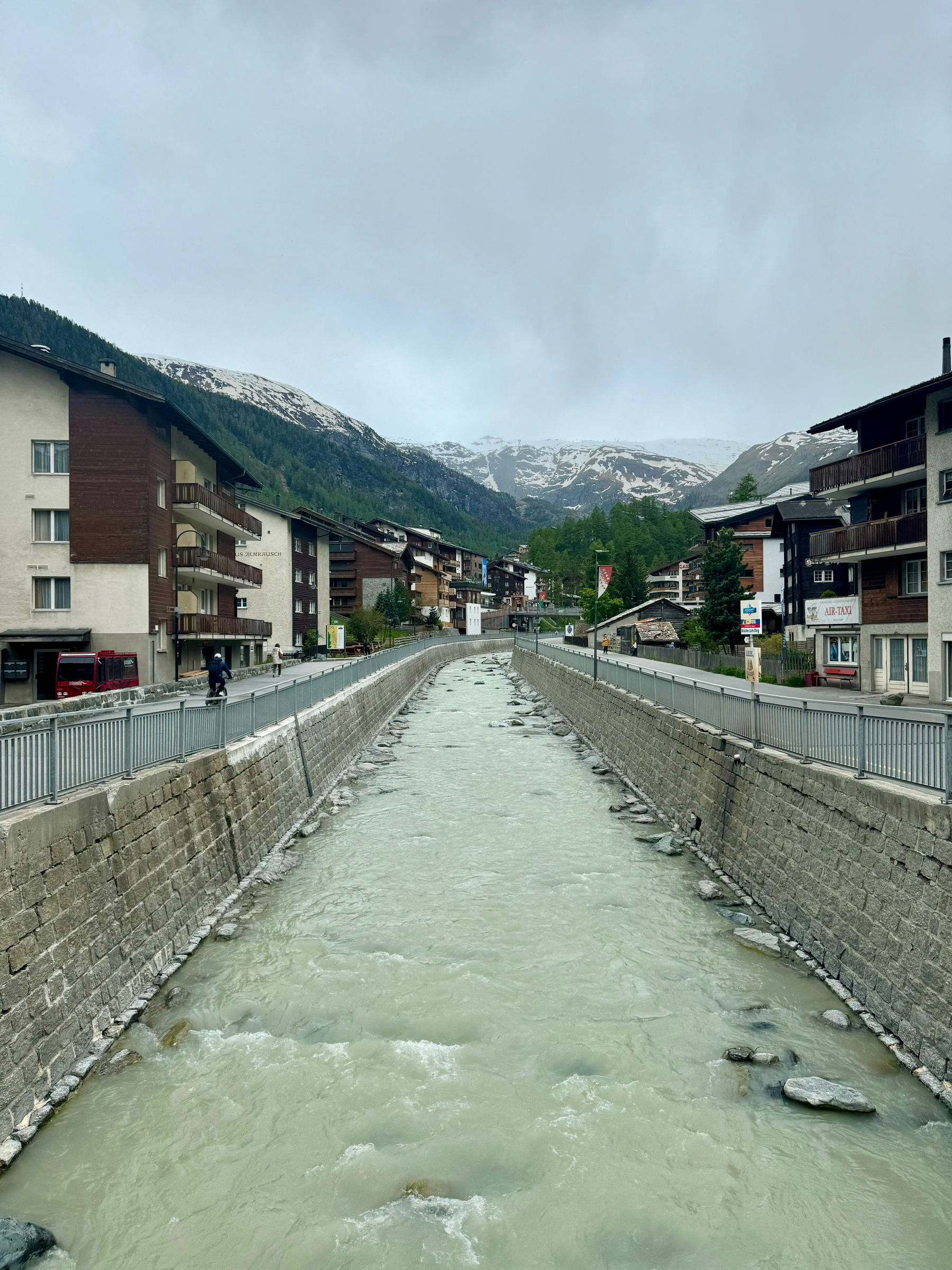 A river flowing through a channel in a mountain town with buildings on either side and snow-capped mountains in the background under a cloudy sky.