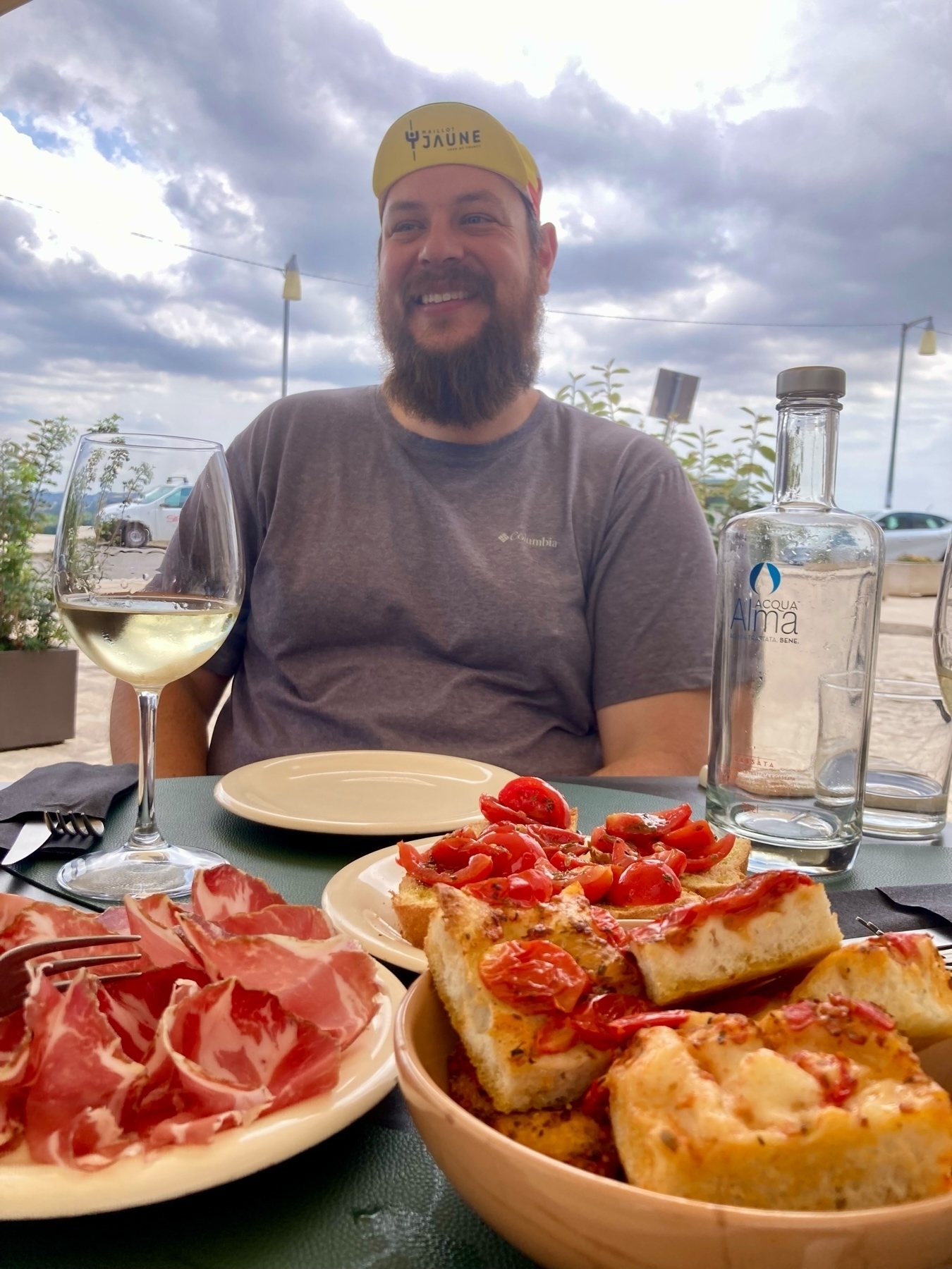 A man with a beard and a yellow cap sits at an outdoor table. In front of him are a glass of white wine, a plate of sliced cured meat, and a bowl of bread topped with cherry tomatoes. 