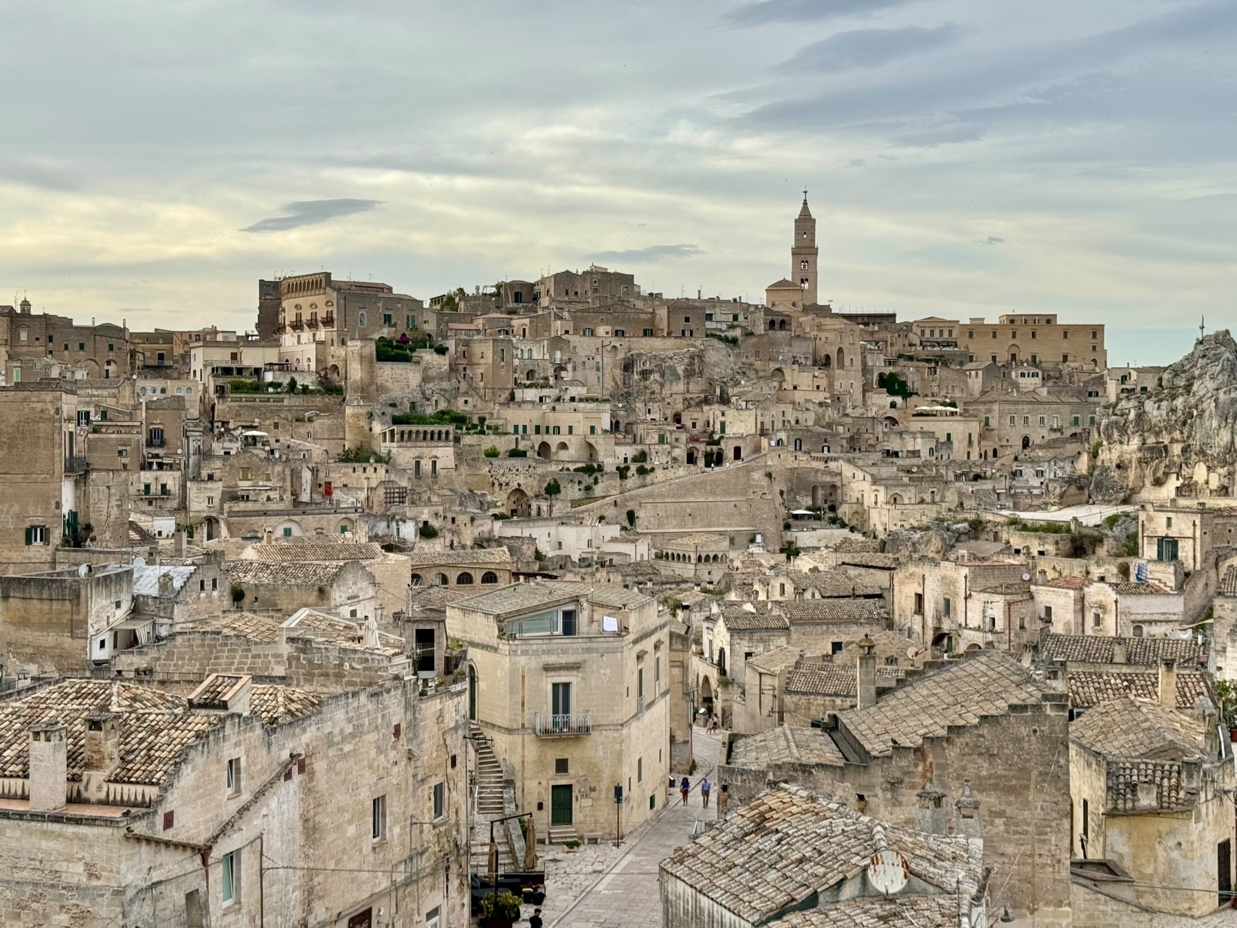 View of the ancient city of Matera in Italy, featuring a dense cluster of historic stone buildings with tiled roofs and narrow streets. The landscape is dominated by a church tower rising above the city. The sky is overcast with scattered clouds.