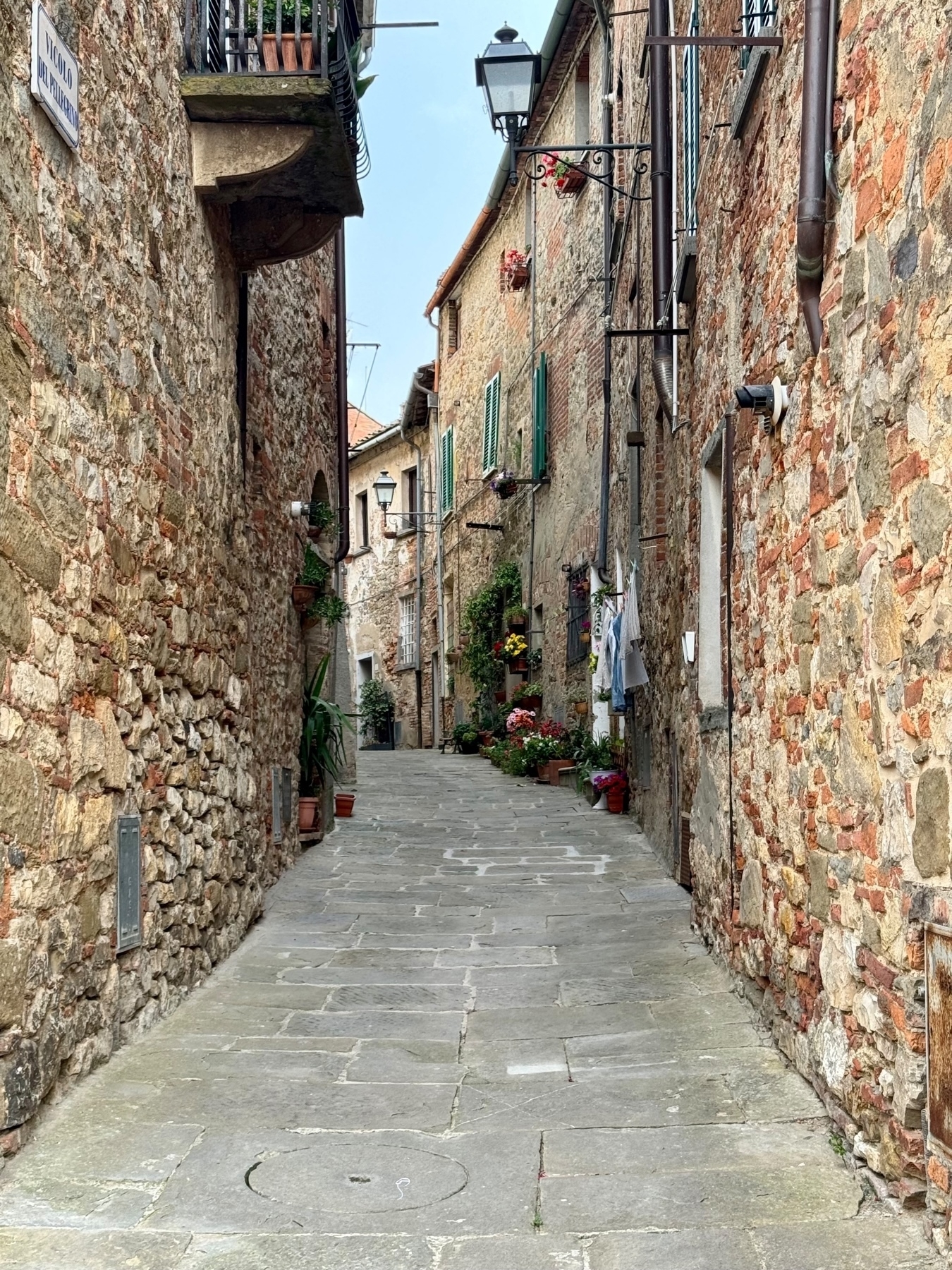 A narrow, stone-paved street in a quaint village or historic town. The street is lined with aged, stone buildings adorned with green window shutters and flower pots. Lamps hang from the sides of buildings. 