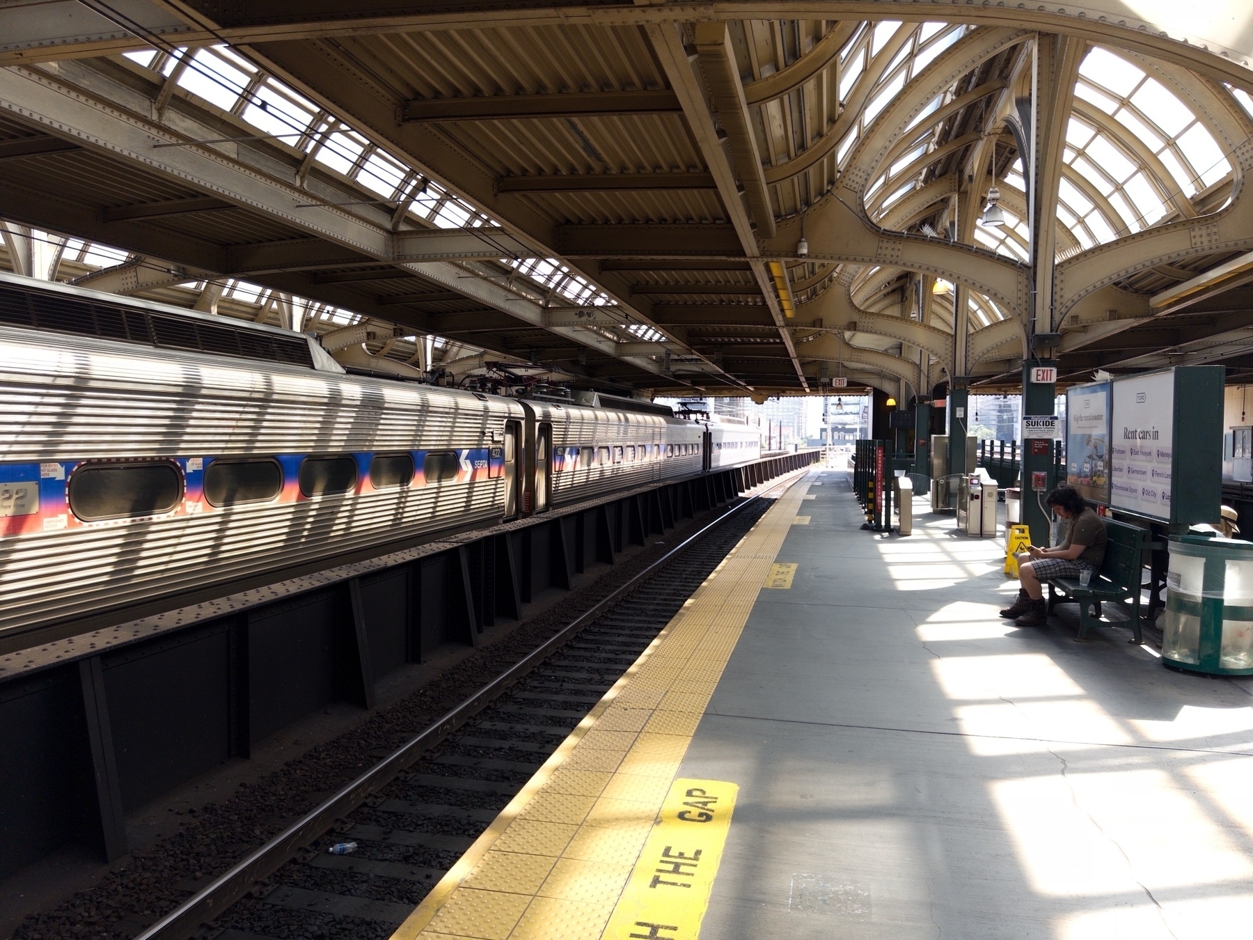 A train station platform featuring a metallic train with blue, white, and red stripe details. The platform has a yellow “WATCH THE GAP” warning strip along the edge. The station has an arched, ornate roof structure with skylights.