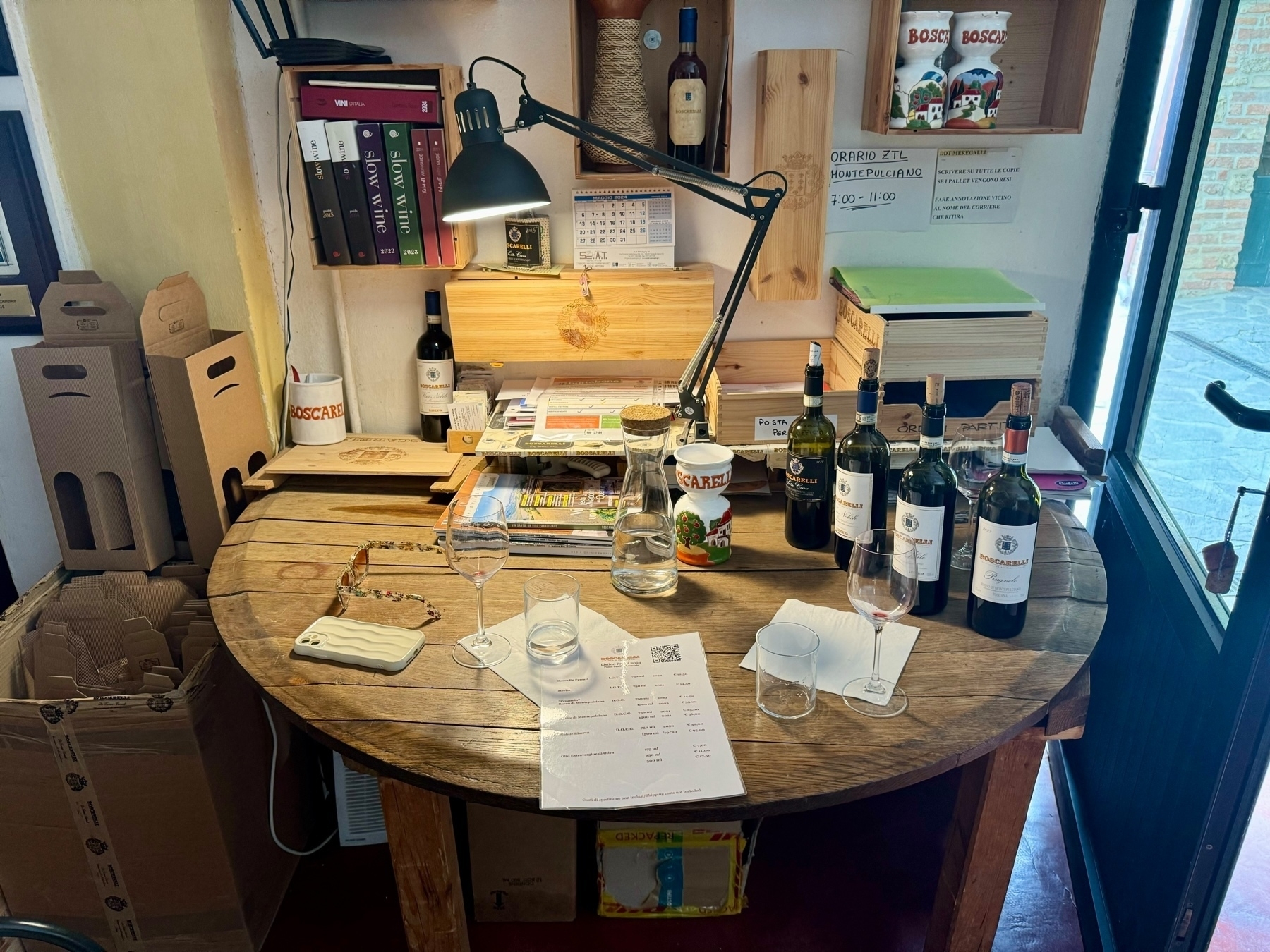 A rustic wooden table with various items, including wine bottles, glasses, papers, and a smartphone case. Shelves and boxes containing books, wine packaging, and other items surround the table. A desk lamp illuminates the area. 