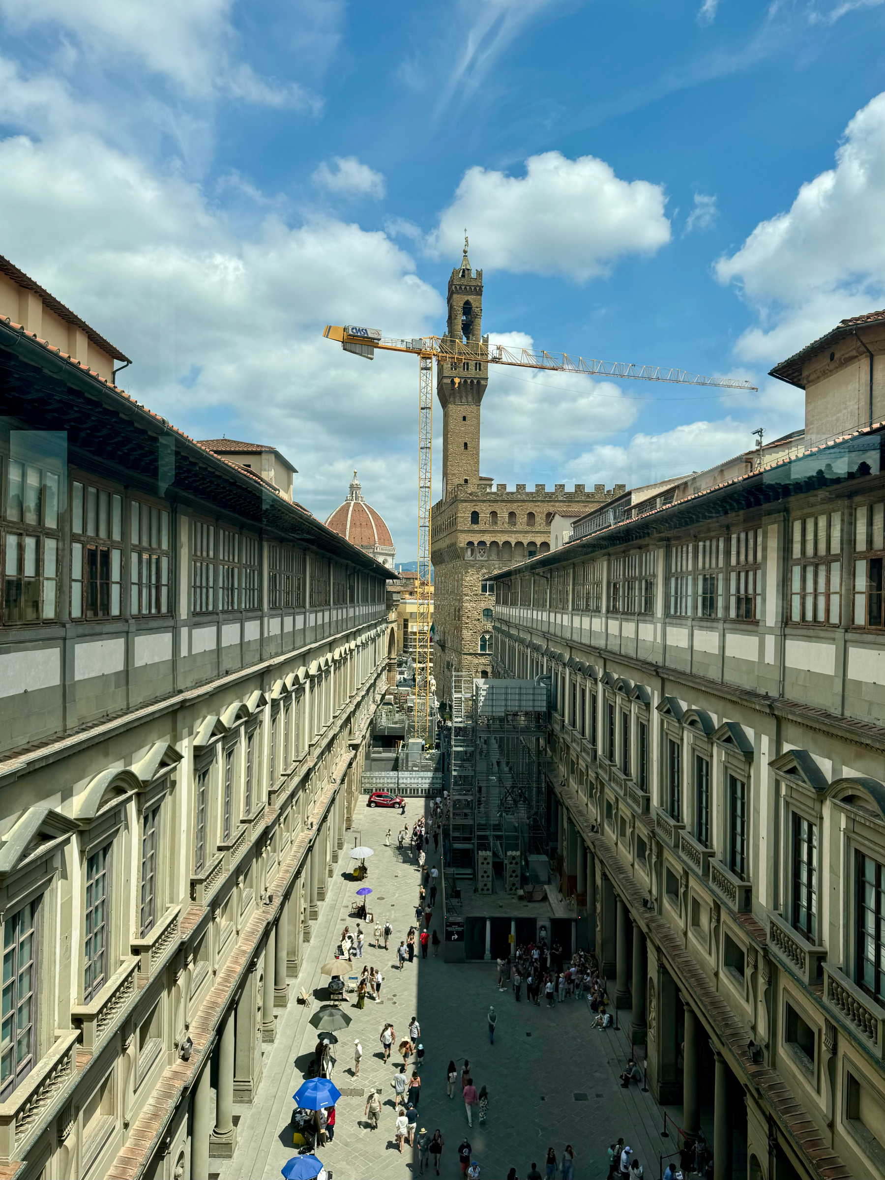 A view of the courtyard of the Uffizi Gallery in Florence, Italy. The street is crowded with people, some holding umbrellas for shade. A prominent bell tower and the dome of the Florence Cathedral can be seen in the background. 