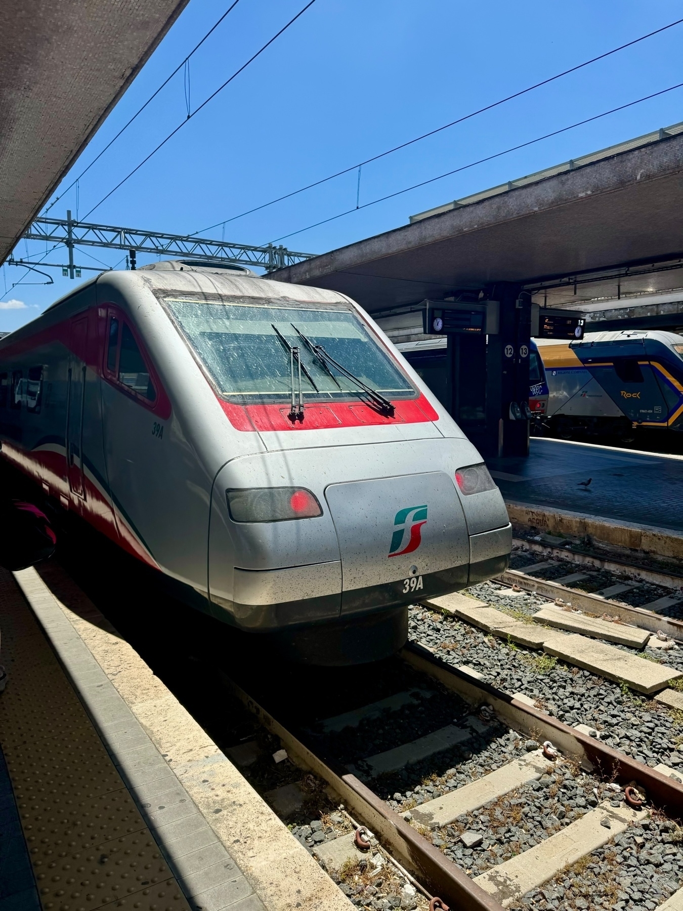 A modern high-speed train on a railway track at a train station, with another train visible in the background. It is a sunny day with clear blue skies, and the station platform is partially shaded. 