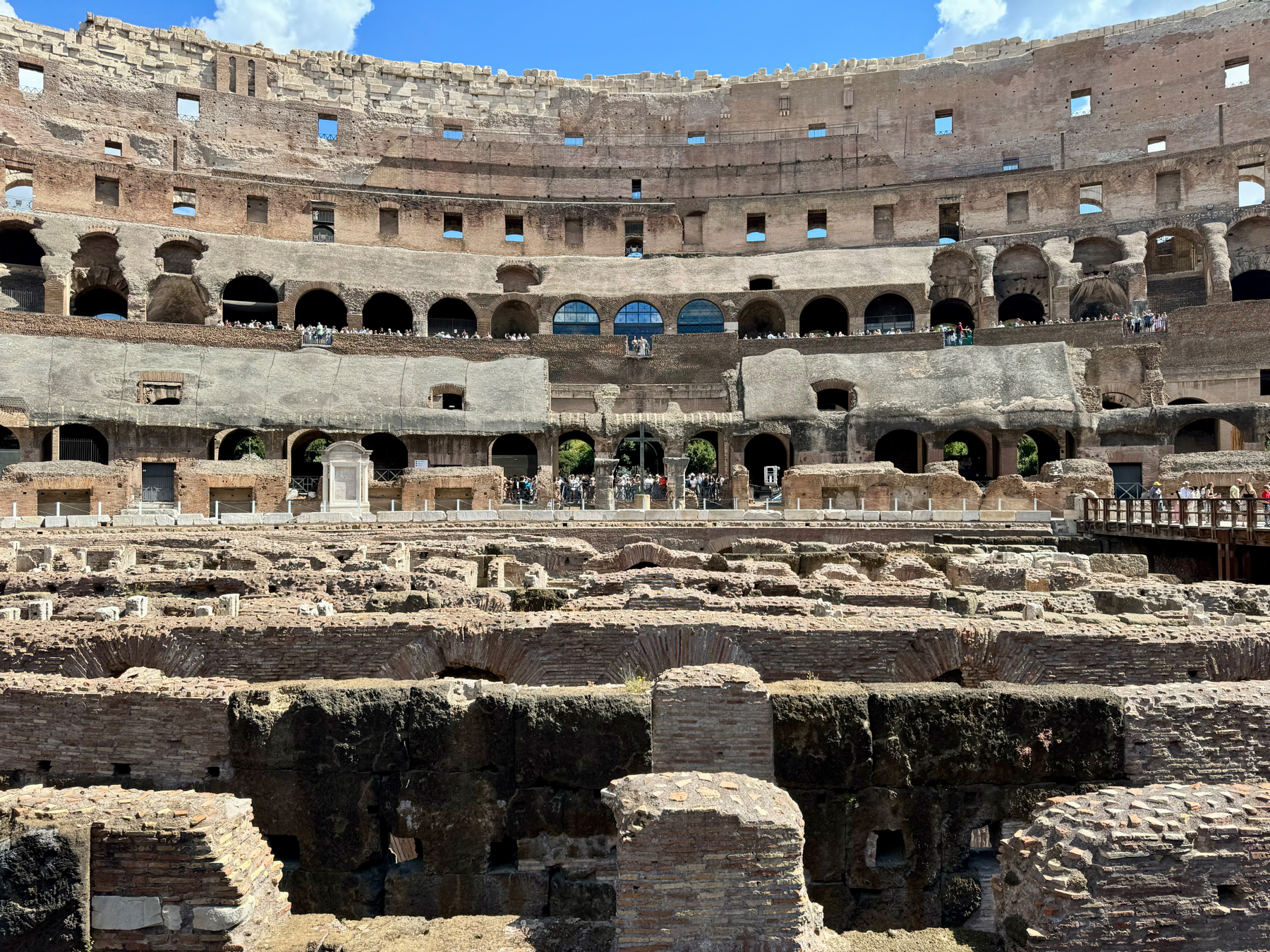 Interior of the Colosseum in Rome, featuring ancient stone ruins and structural remains of the amphitheater. Multiple levels of arches and seating are visible, alongside groups of visitors exploring the site. Bright sunny sky above with minimal clouds.