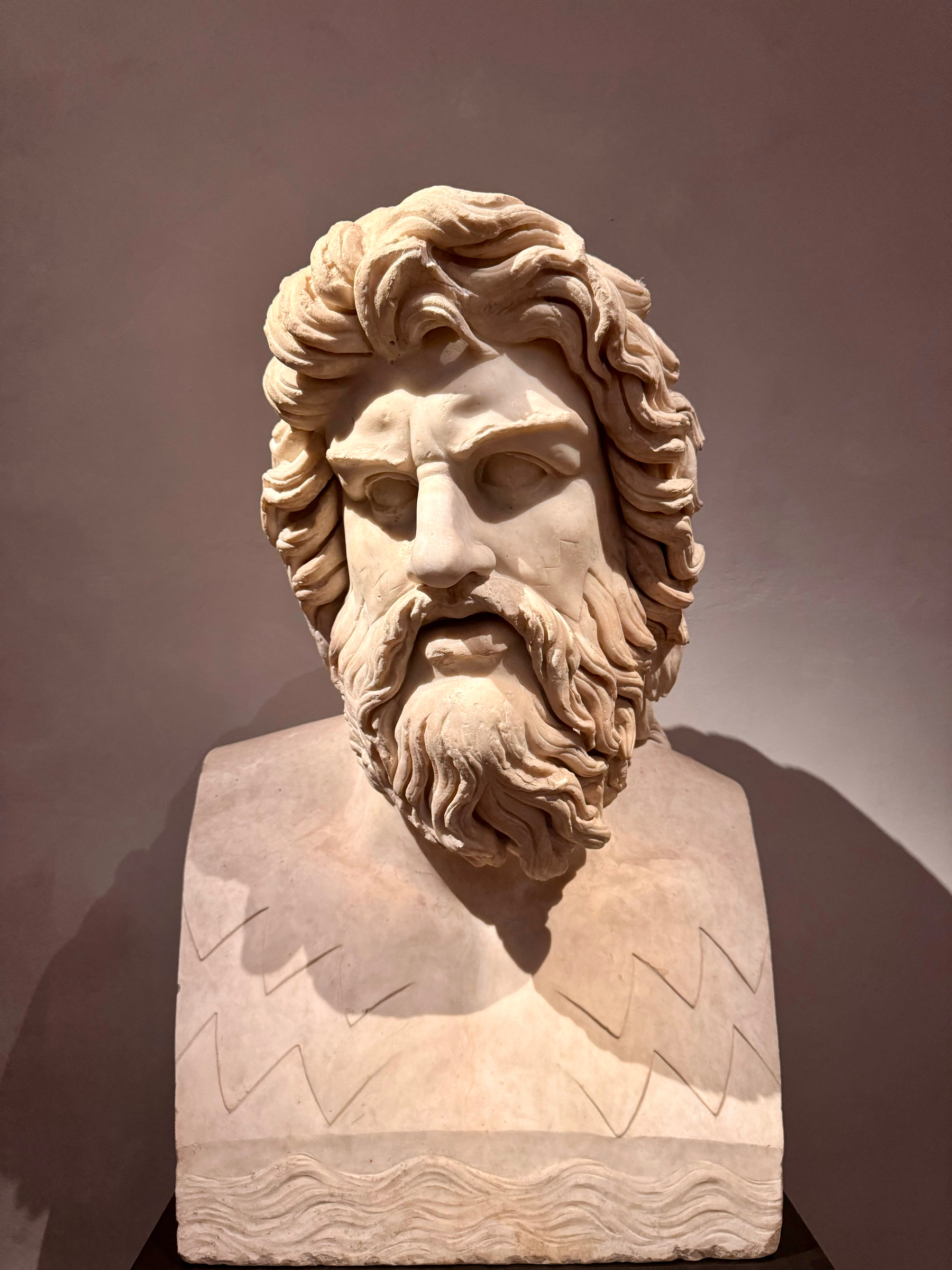 A marble bust of a bearded man with flowing hair, depicted with a stern expression. Carved waves and zig-zag patterns are present on the bust’s base. The sculpture is set against a plain background.