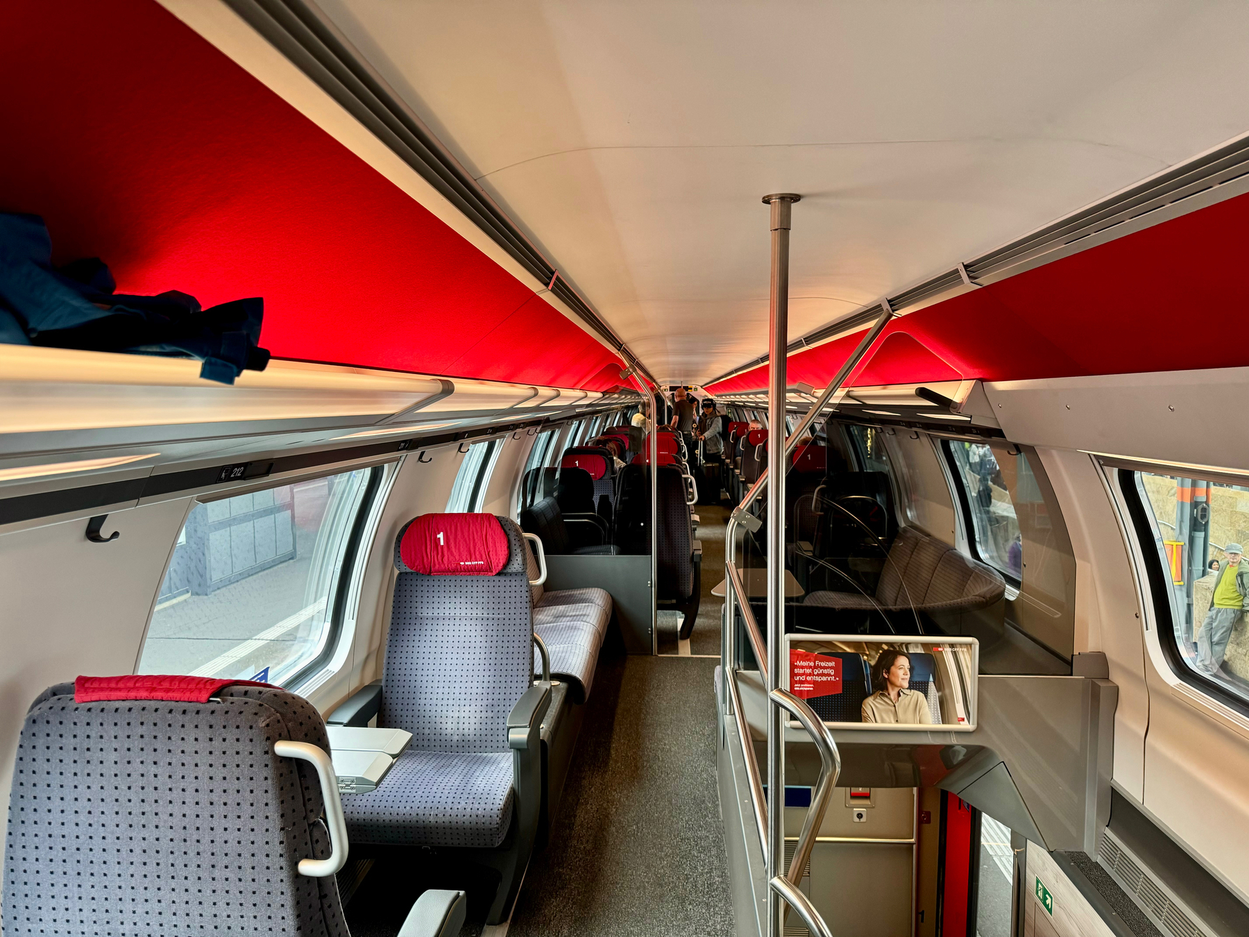 Interior of a modern train carriage with red and grey upholstery, overhead storage, and large windows. A television screen shows an advertisement. Passengers are by the door, and a worker is visible through the window.