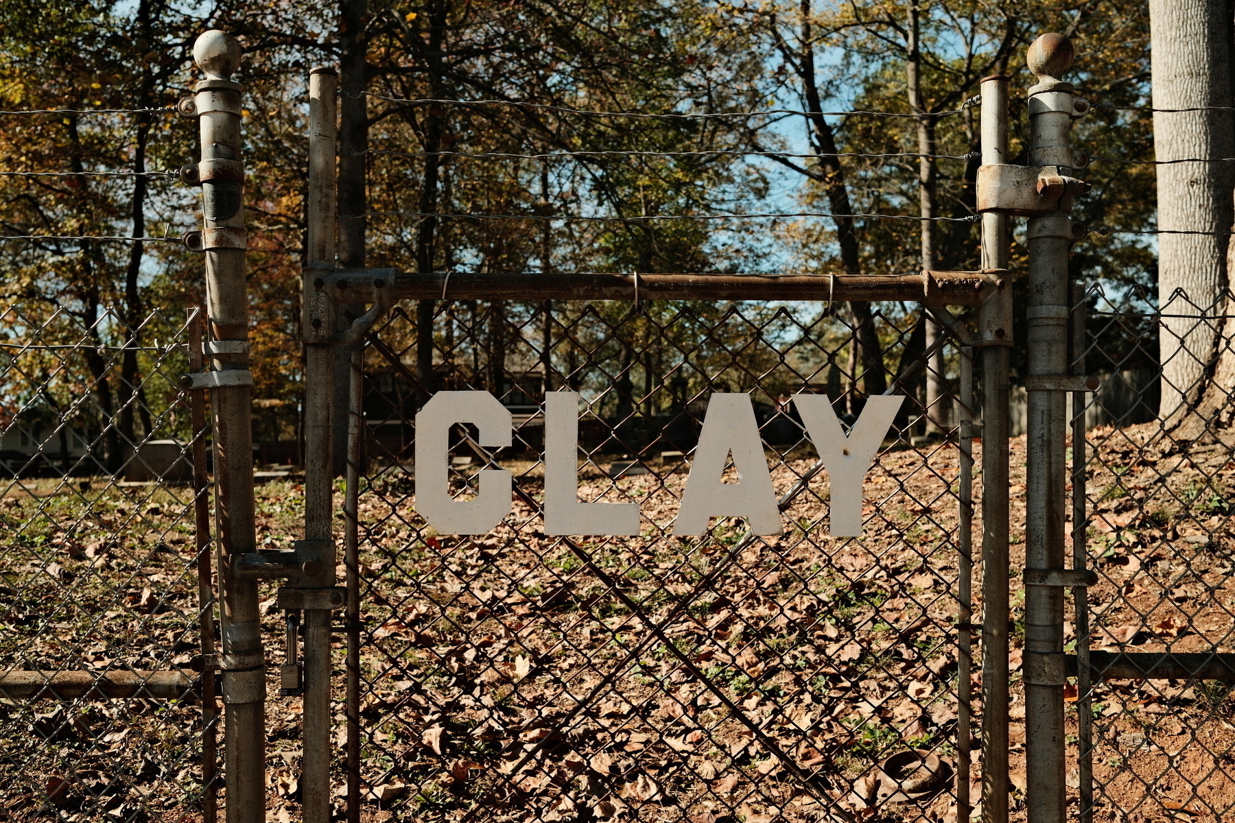 A rusty chain-link gate with the word CLAY in large letters stands amidst fallen leaves, trees, and cemetary in the background.