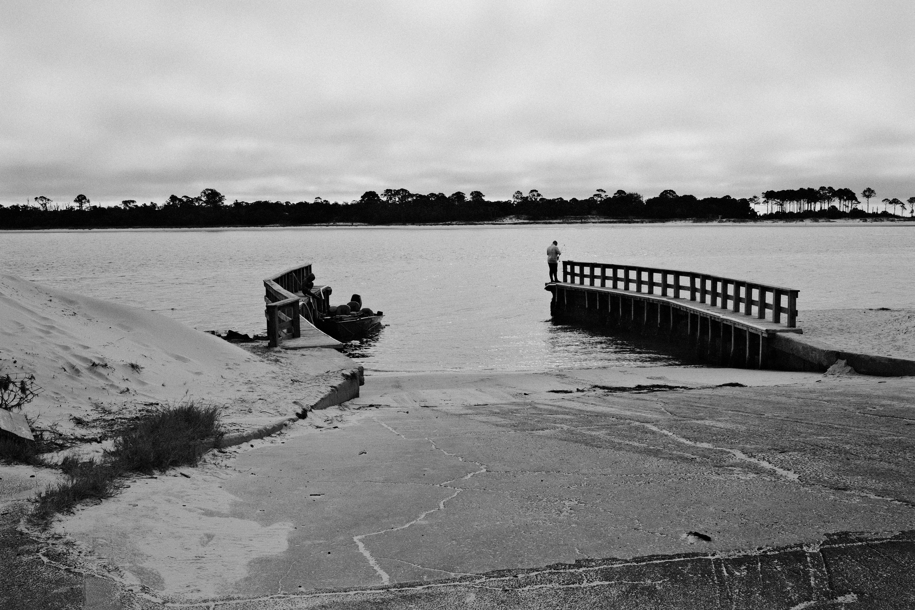 Auto-generated description: A person stands on a wooden dock overlooking a body of water, with another individual sitting by a small beached boat under a cloudy sky.