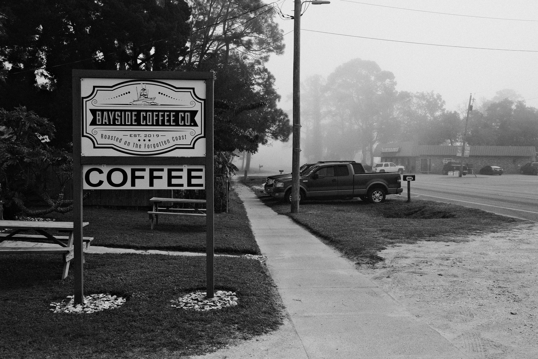 Auto-generated description: A black and white photo depicts a sign for Bayside Coffee Co. next to a roadside with parked vehicles and trees in the background.