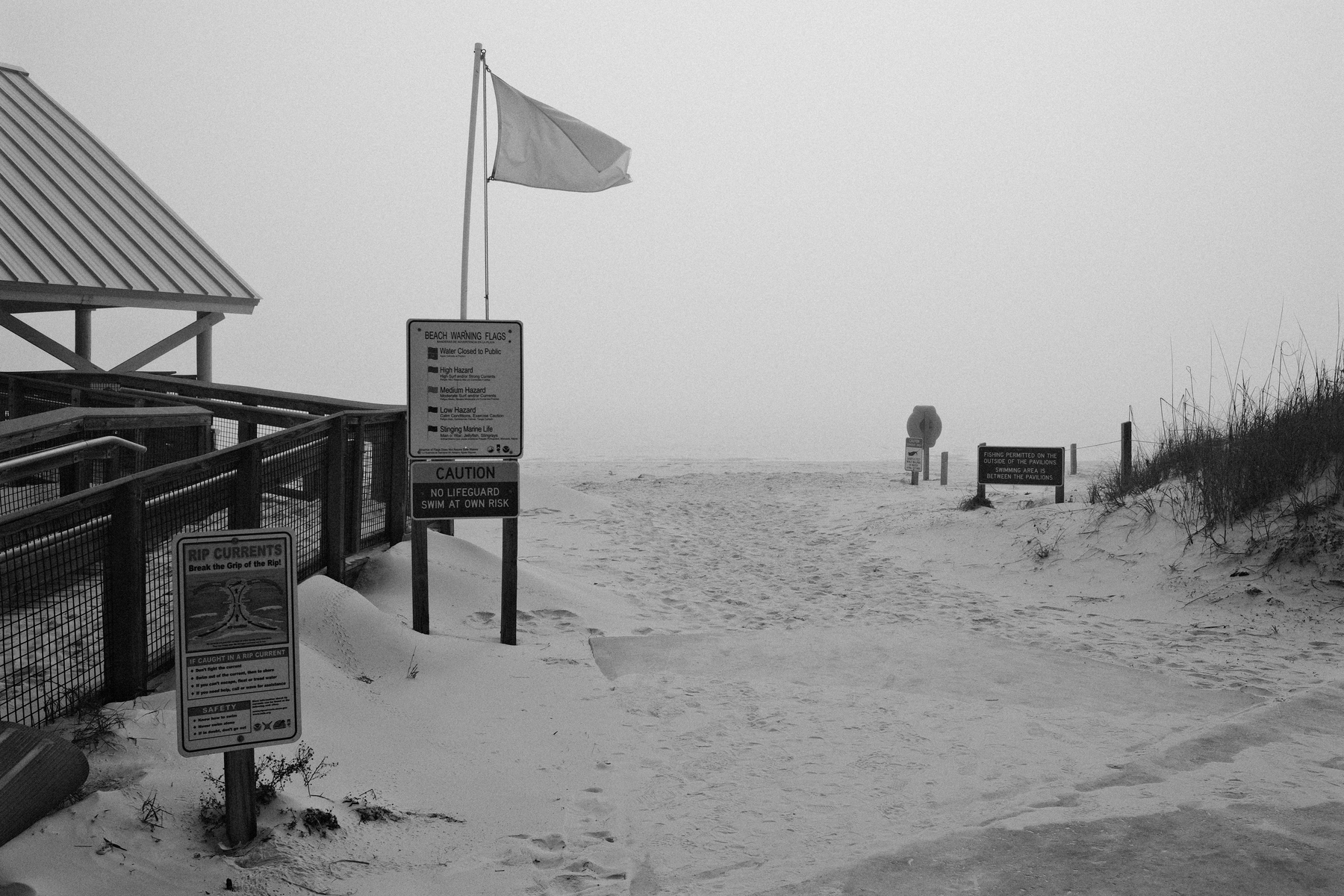 Auto-generated description: A foggy beach scene features a lone figure in the distance, warning signs, and a flapping flag near a fenced walkway.