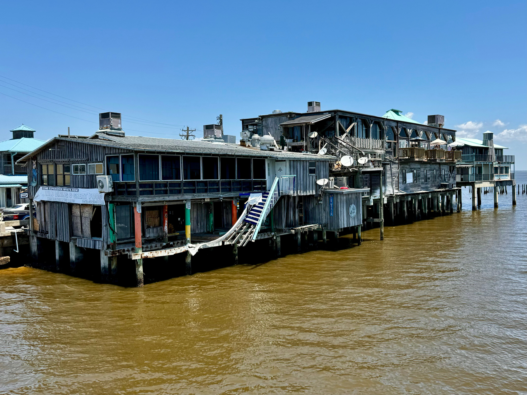 Wooden buildings on stilts over a muddy body of water, featuring balconies, stairs, and various satellite dishes and air conditioning units.