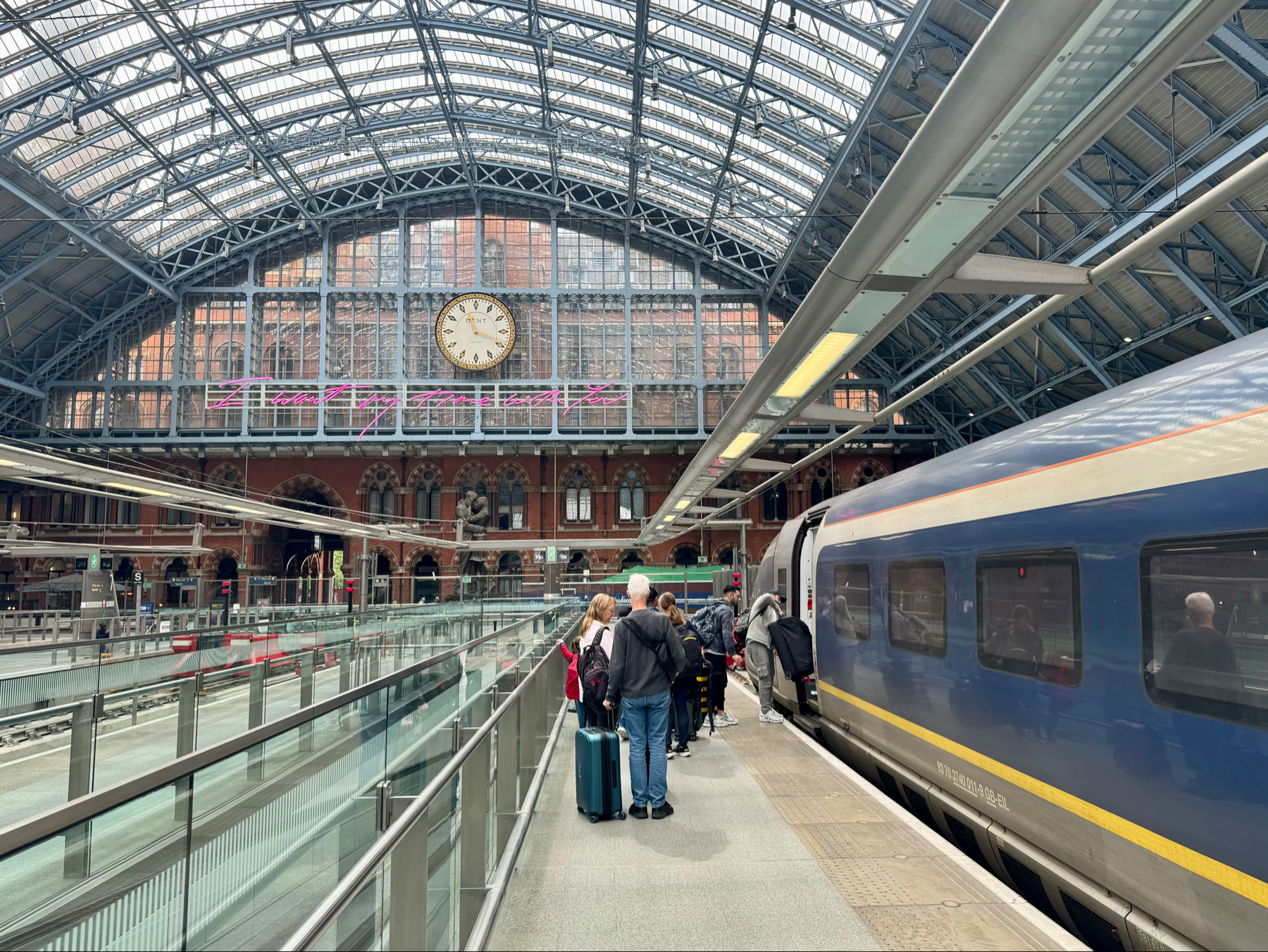 A busy train station with a large clock, passengers with luggage, a departing train, and a glass and metal roof structure.