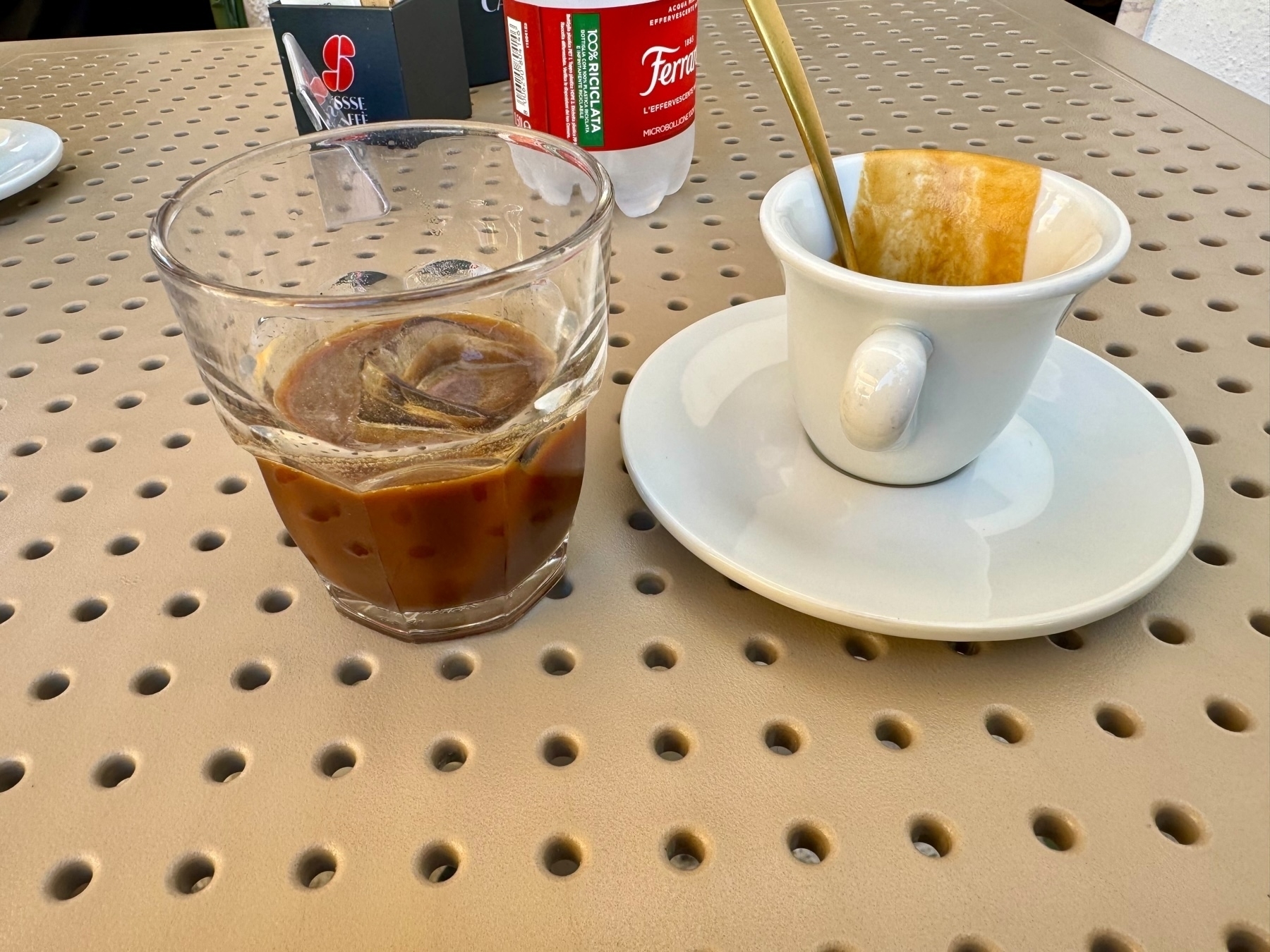 A glass of iced coffee sits next to an empty white coffee cup with a spoon inside on a perforated beige table. A small bottle of water and a box of sugar packets are in the background.