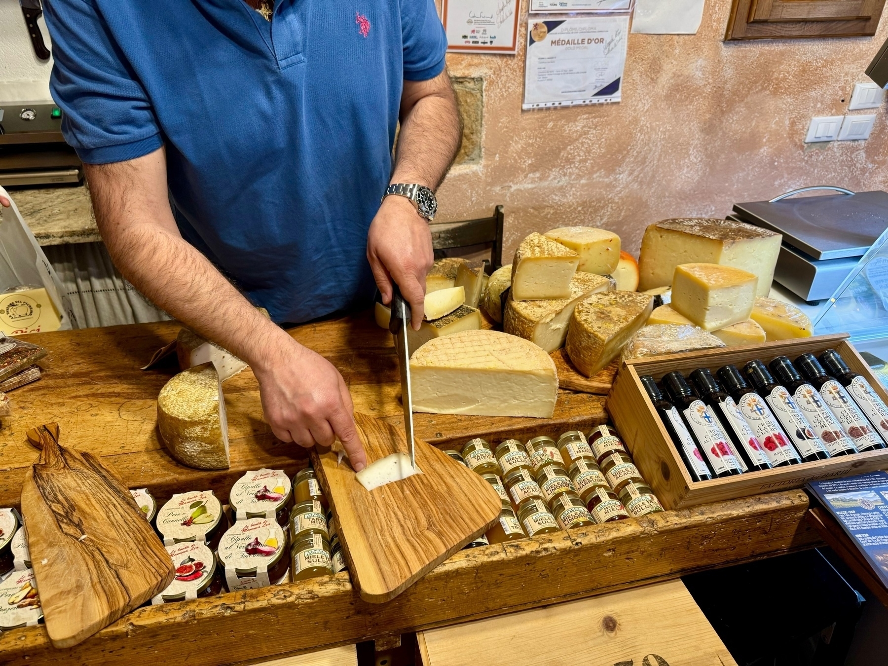 A person in a blue shirt cuts a slice of cheese on a wooden board in a cheese shop. Various types of cheese, small jars of condiments, and bottles of balsamic vinegar are displayed on the wooden counter. 
