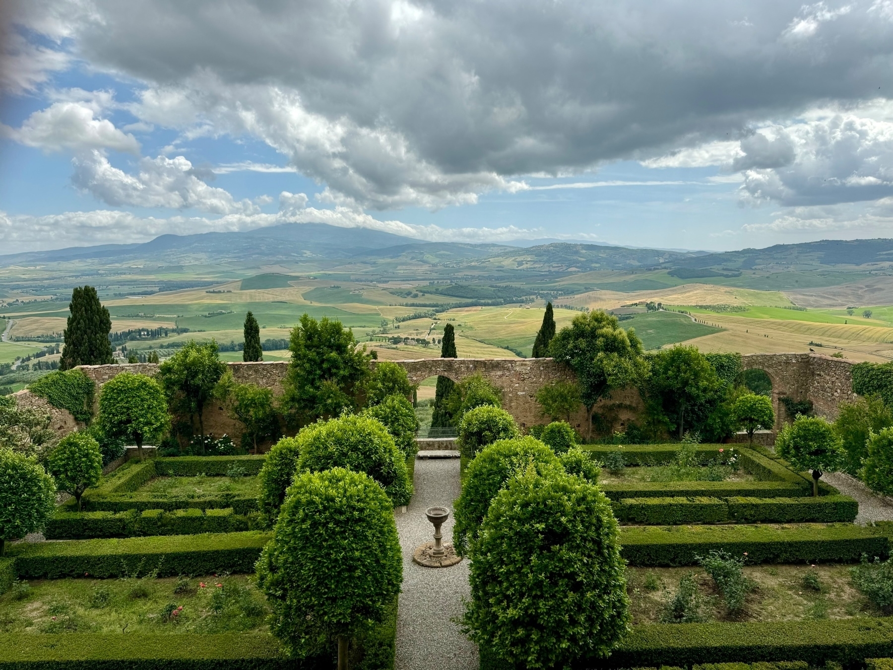 A picturesque landscape featuring a well-manicured formal garden with neatly trimmed bushes and hedges. Beyond the garden, there is an old stone wall partially overgrown with greenery. The backdrop showcases rolling hills, fields, and distant mountains. 