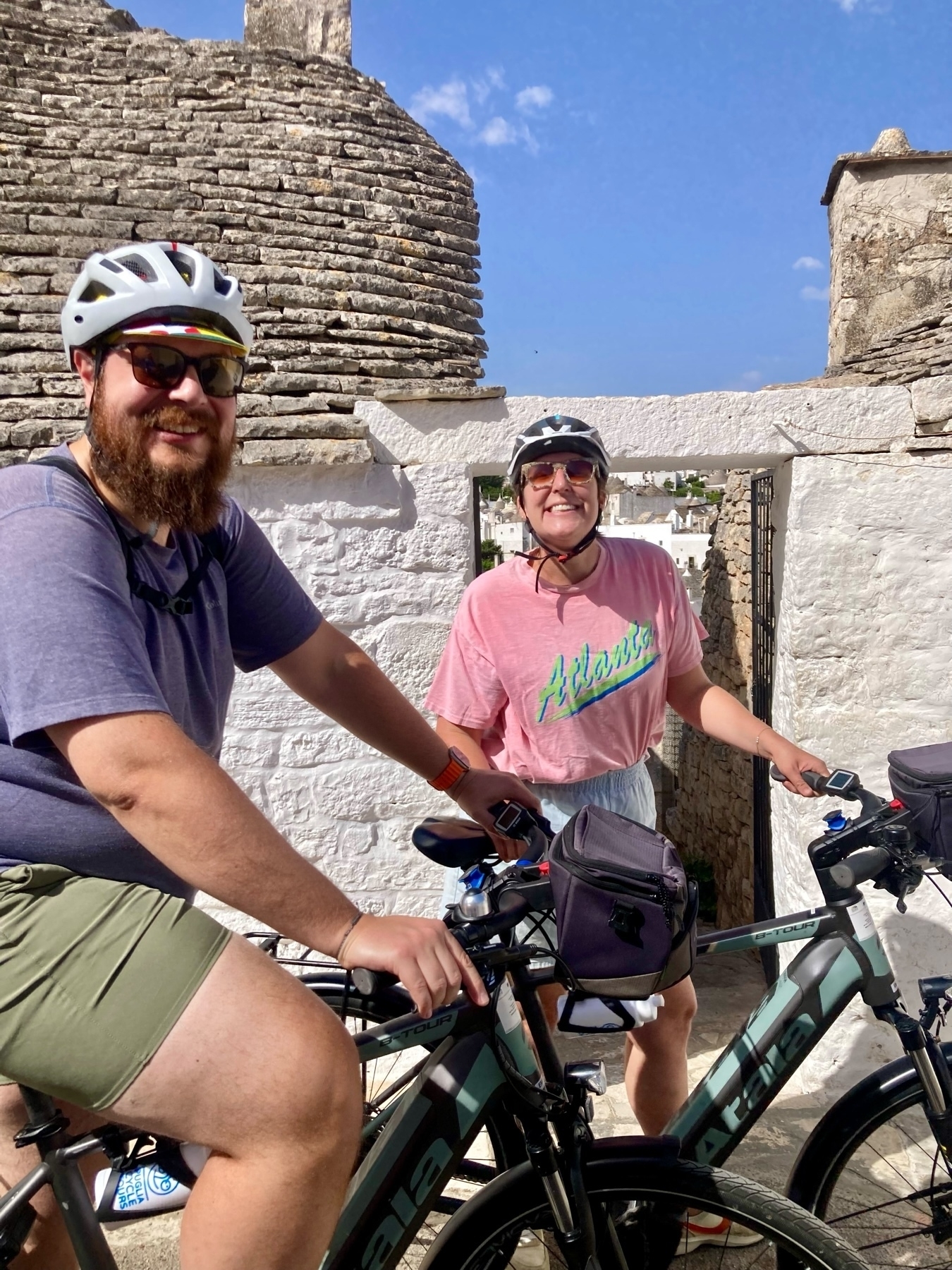 A bearded man and a woman, both wearing helmets and sunglasses, are posing with their bicycles in front of a traditional stone building with a conical roof.