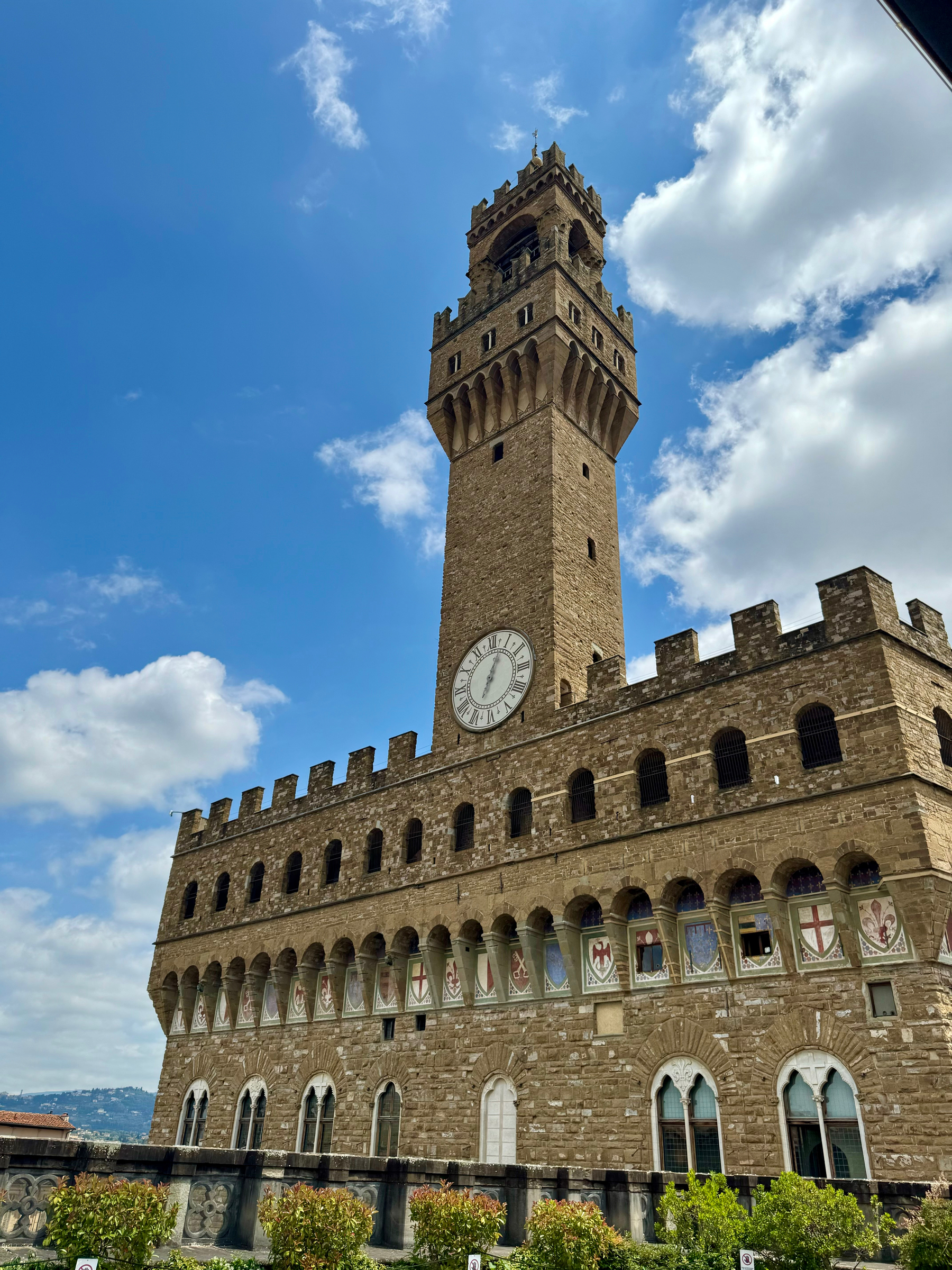 The image depicts the Palazzo Vecchio, a historic palace located in Florence, Italy. The photo showcases the building’s tall stone brick tower with a clock and battlements, set against a backdrop of a blue sky with some clouds.