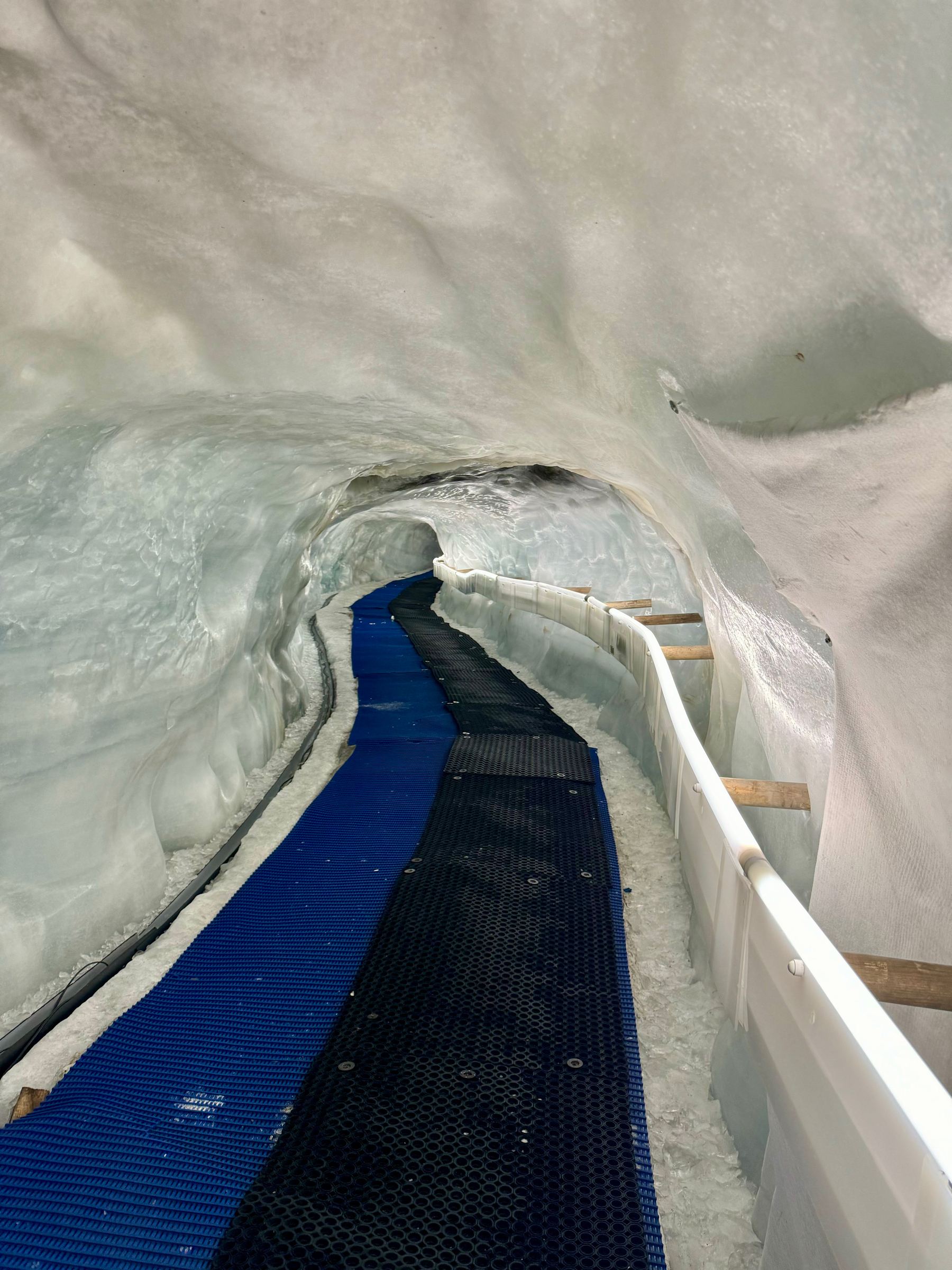 A walkway through an ice tunnel with blue flooring, handrails on the sides, and walls and ceiling of curved, smooth ice.
