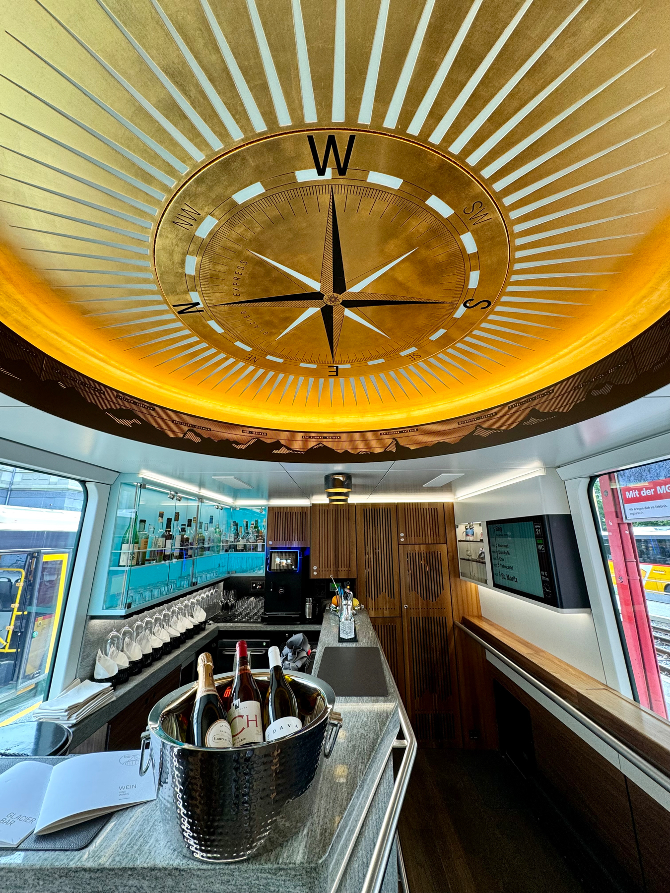 A train bar carriage with a compass rose on the ceiling, a counter with bottles, and a window showing another train outside.