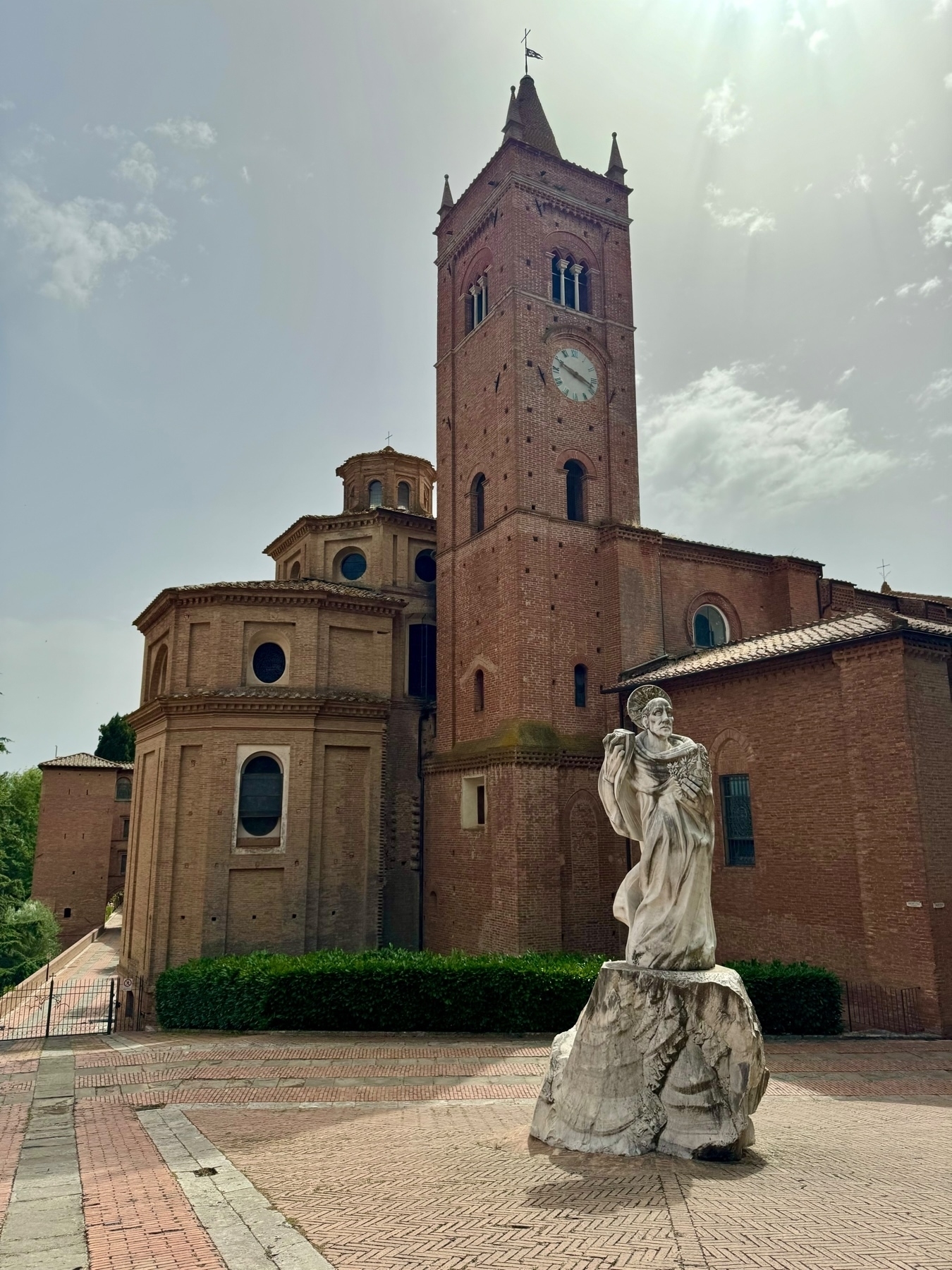 A tall brick church with a clock tower and arched windows under a sunny sky. In front of the church, a white marble statue depicts a robed figure standing on a stone pedestal. The area around the church and statue is paved with red bricks. 